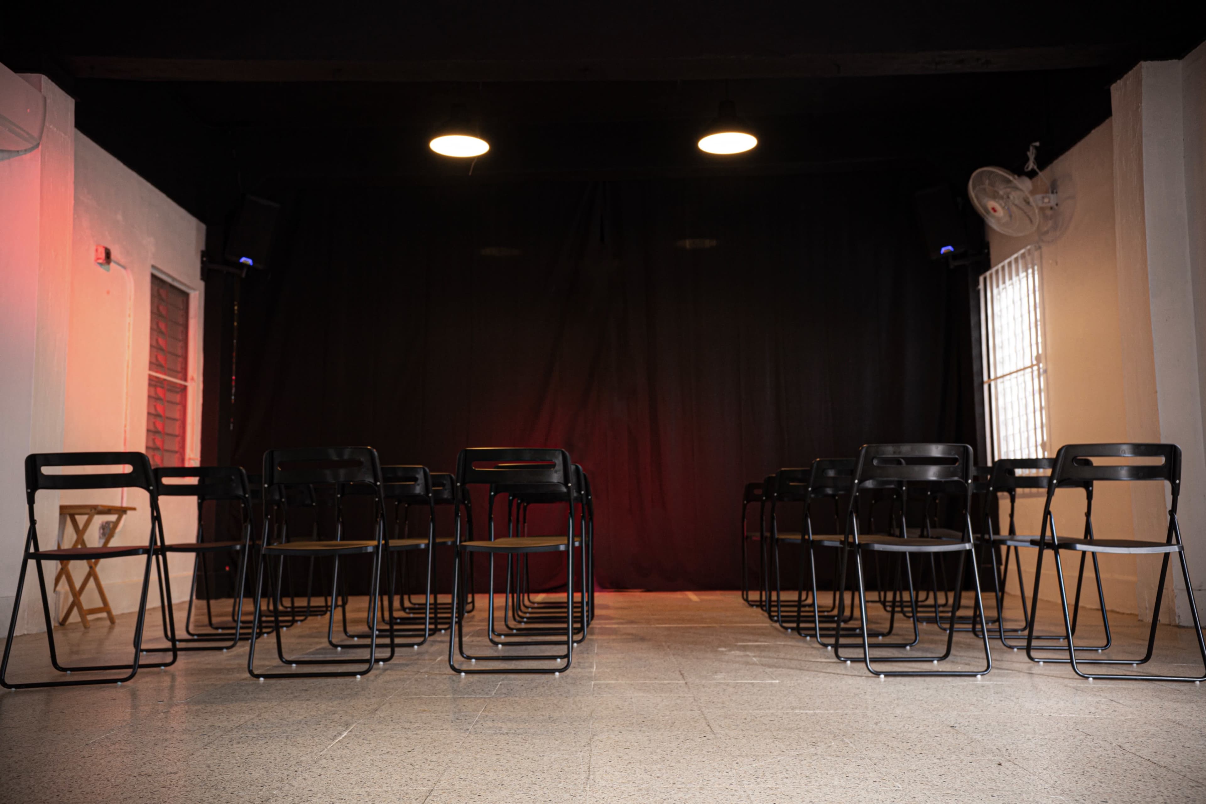 The image shows a small stage area with black chairs arranged in rows facing a dark curtain.