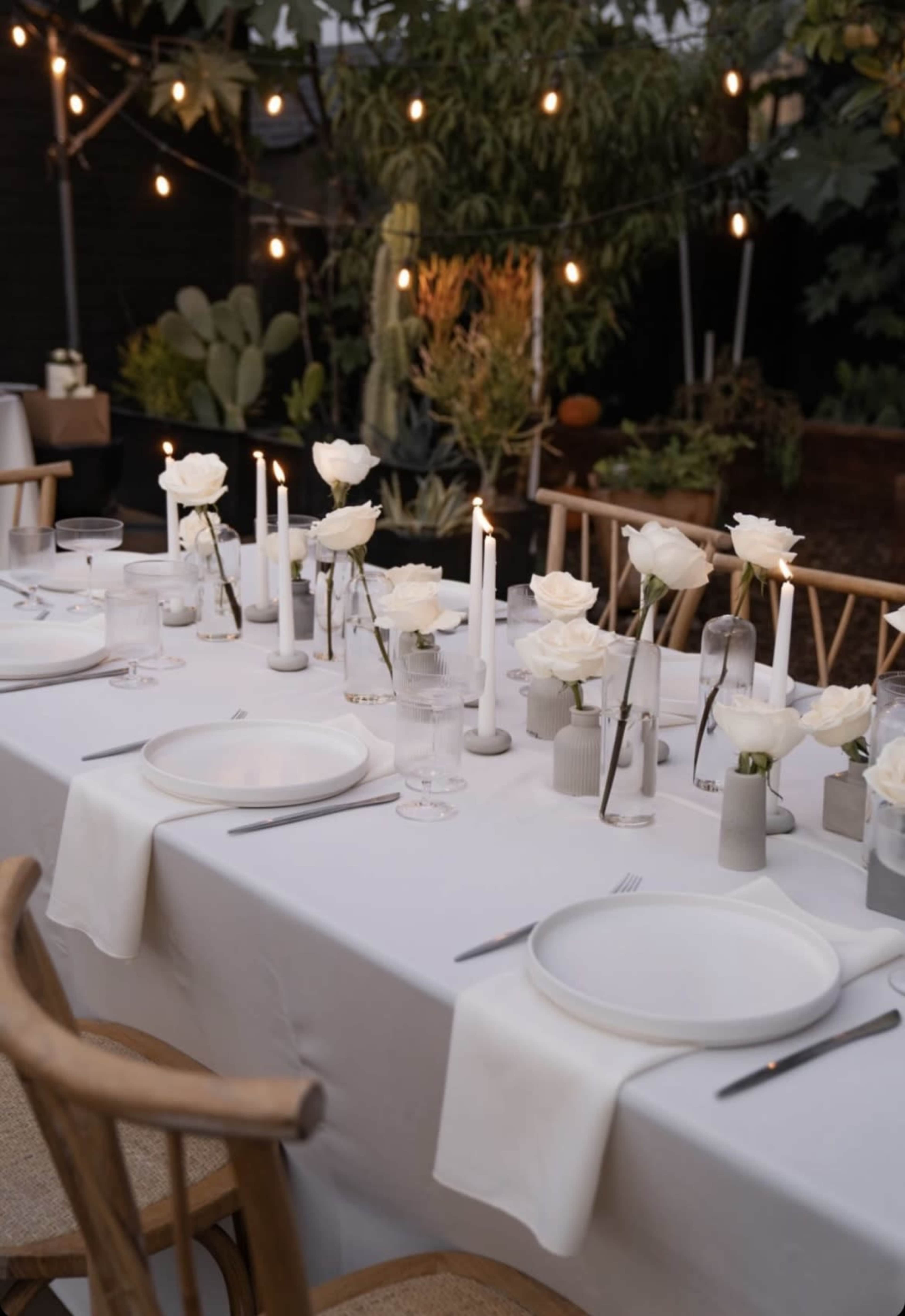 A long dining table is set with white plates, glassware, and vases of white roses, surrounded by plants and illuminated by string lights.