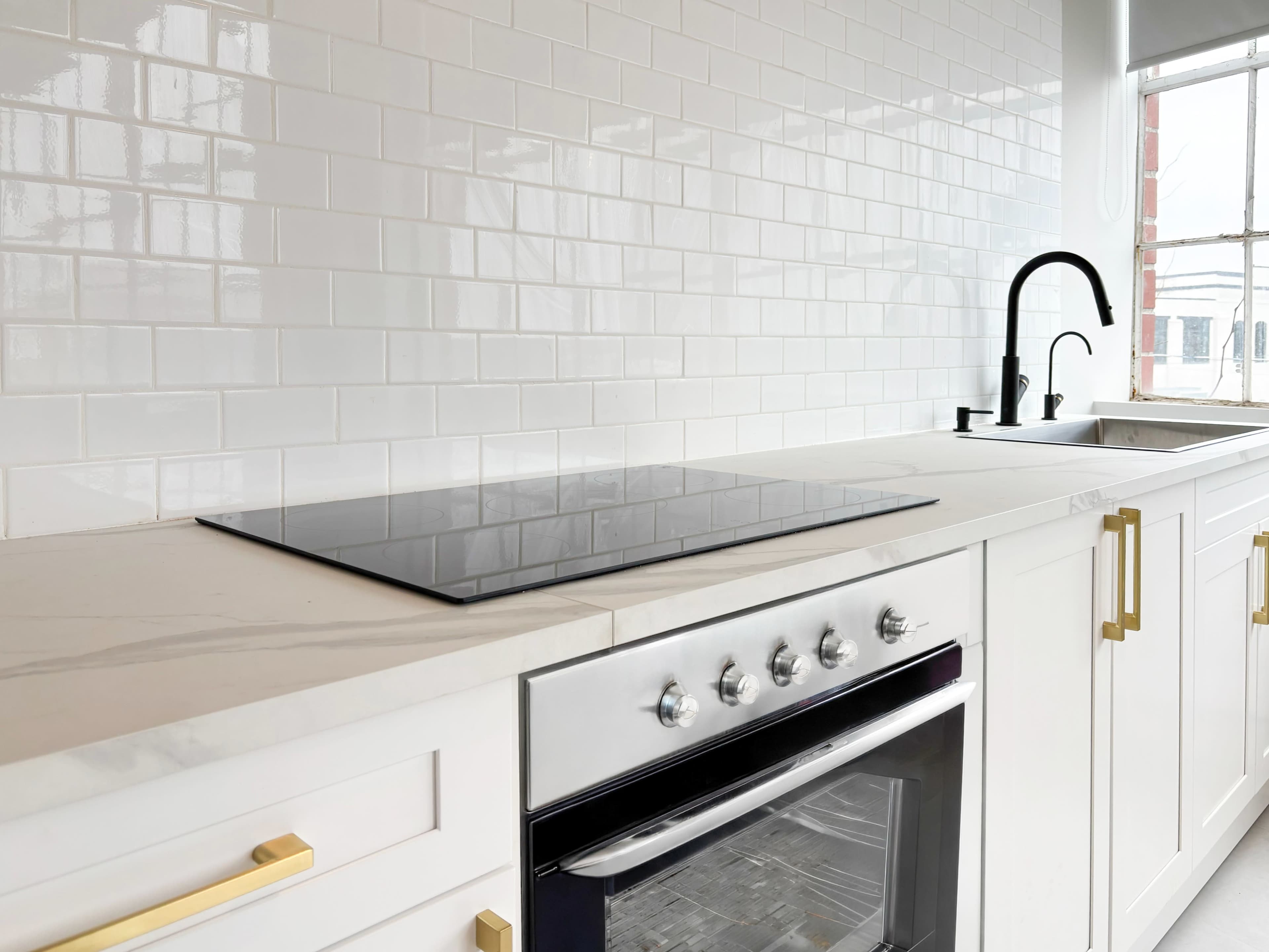 The image features a modern kitchen with a sleek white backsplash, a black cooktop on the countertop, and a stainless steel oven beneath it.