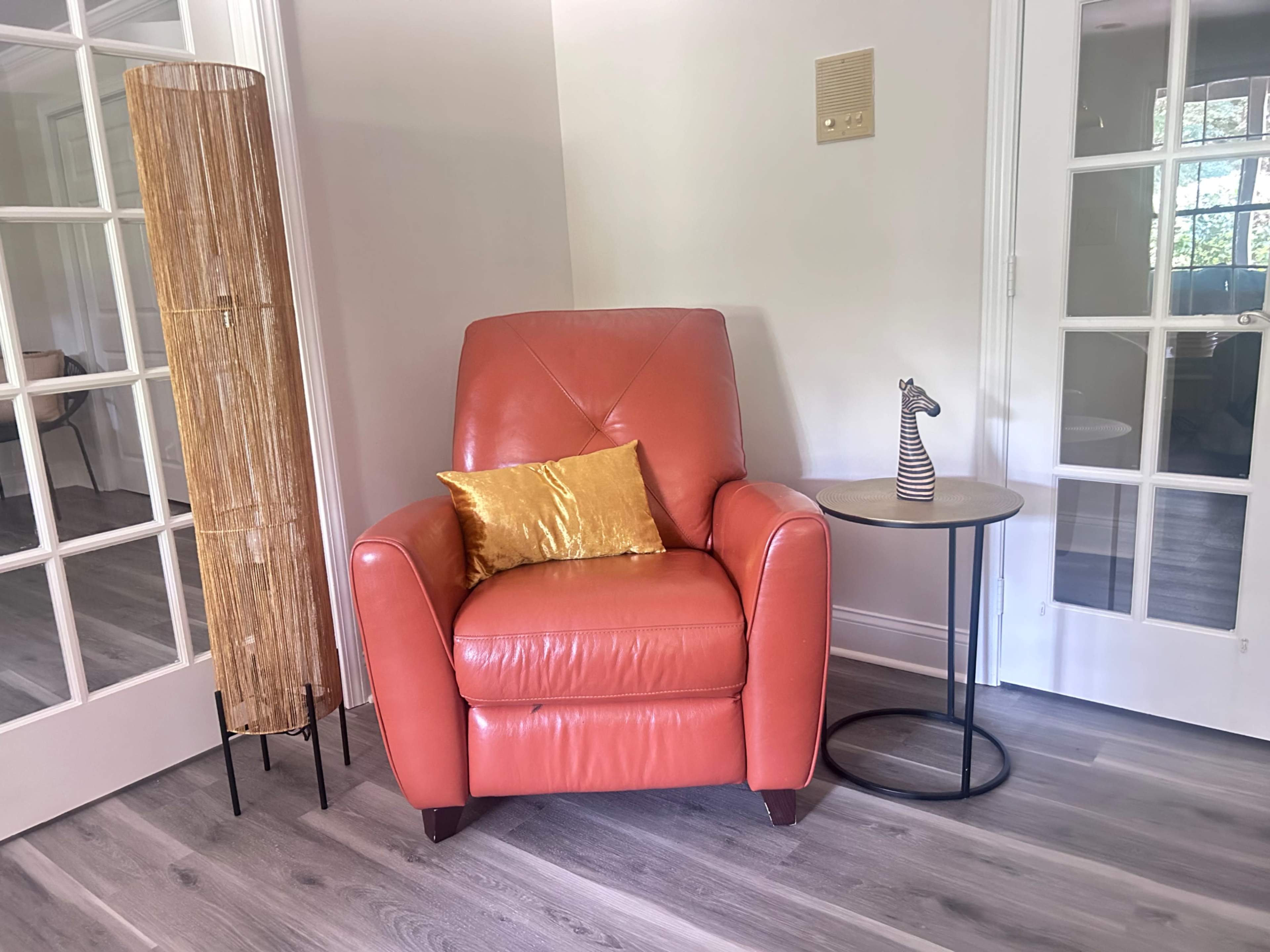 A coral-colored leather armchair is situated next to a small round table and a tall, textured lamp in a bright room.