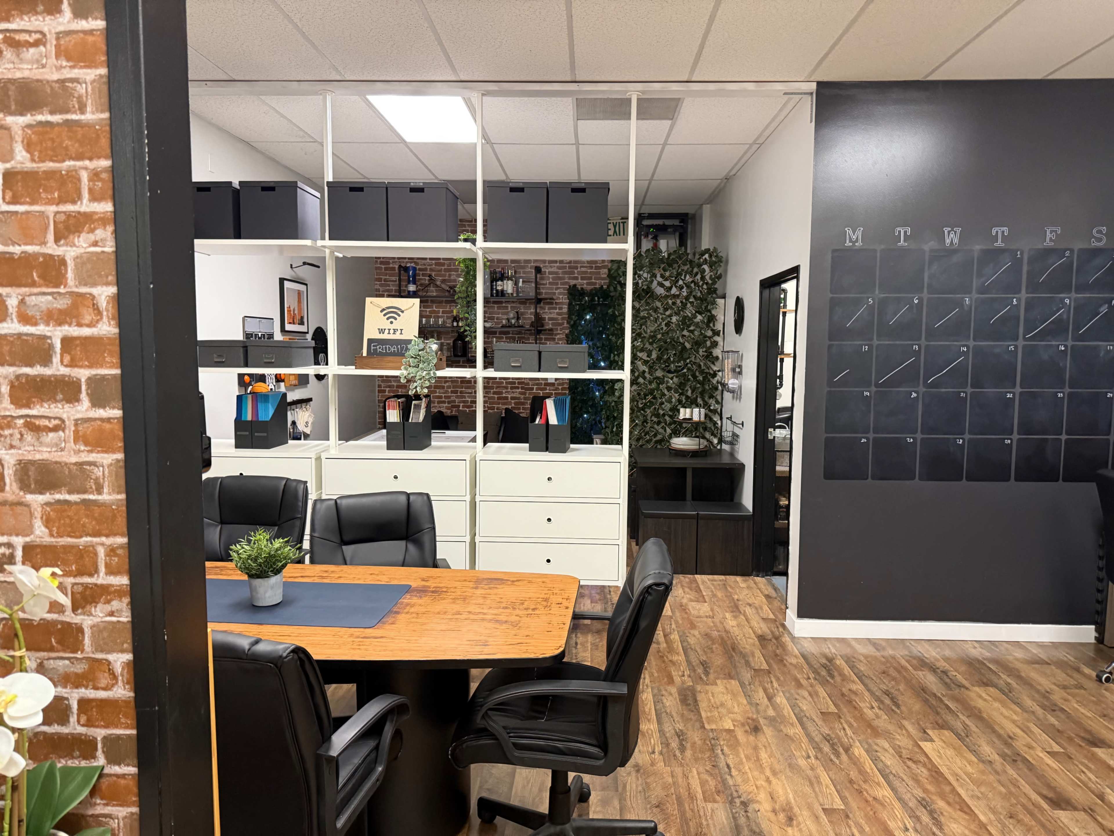 The image shows an office space with a wooden conference table, black chairs, and shelves organized with boxes and decorative plants against a brick wall.