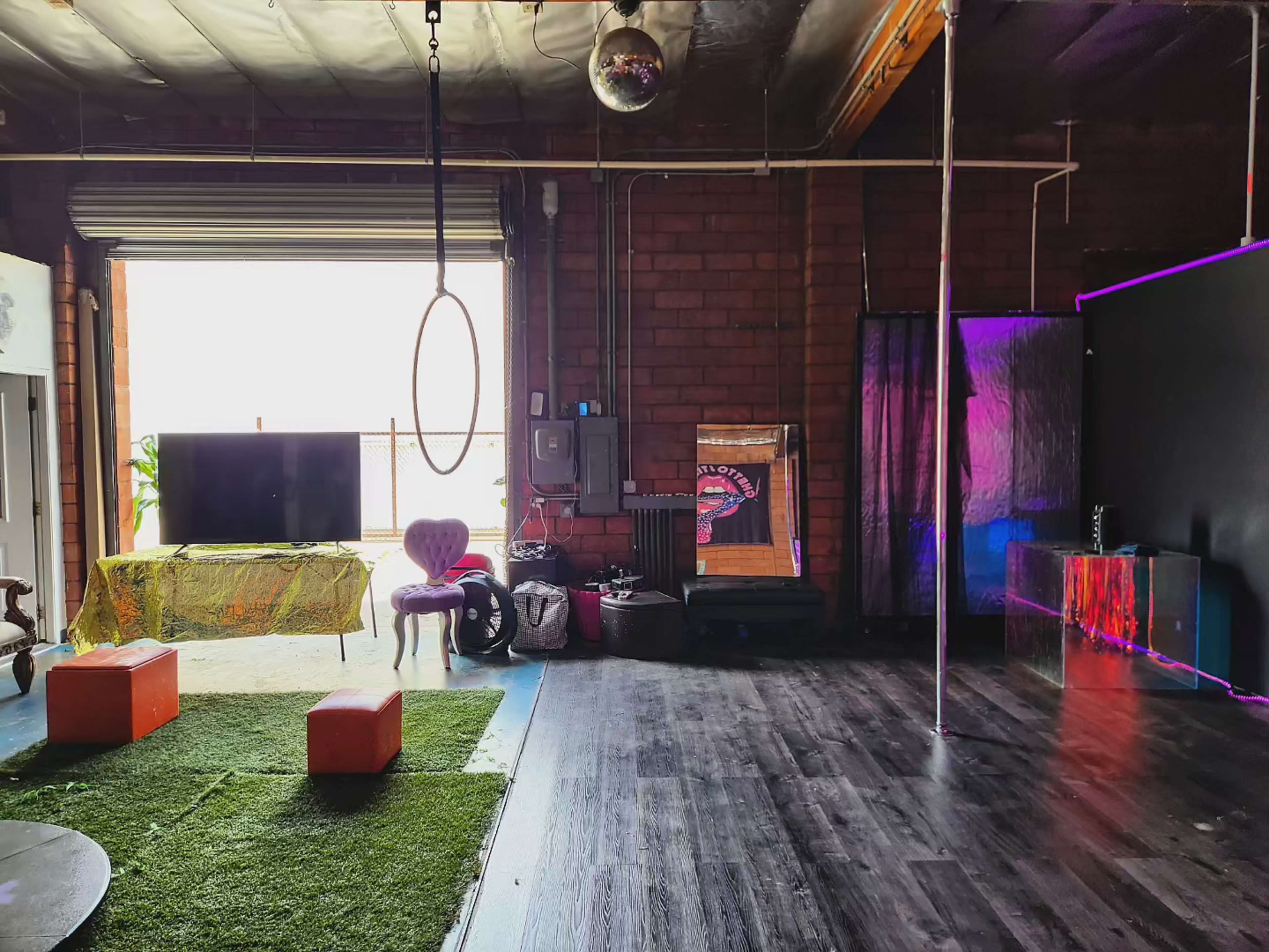 The image shows an indoor space featuring a pole, a hoop hanging from the ceiling, a green carpet area, a television, and various seating arrangements.