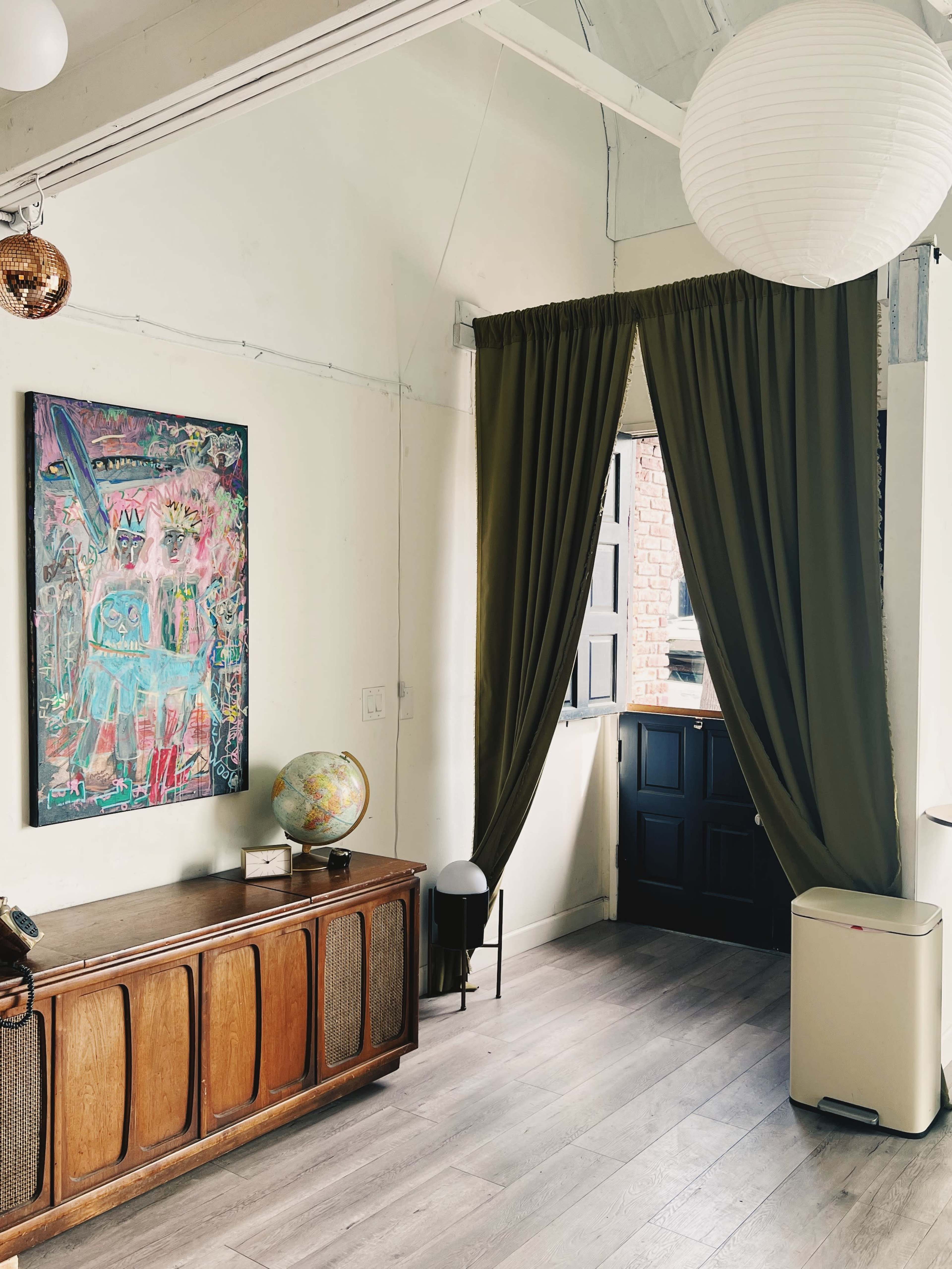 The image shows a bright room featuring a colorful painting on the wall, a wooden cabinet, a globe on a side table, and a dark door partially opened beside a green curtain.