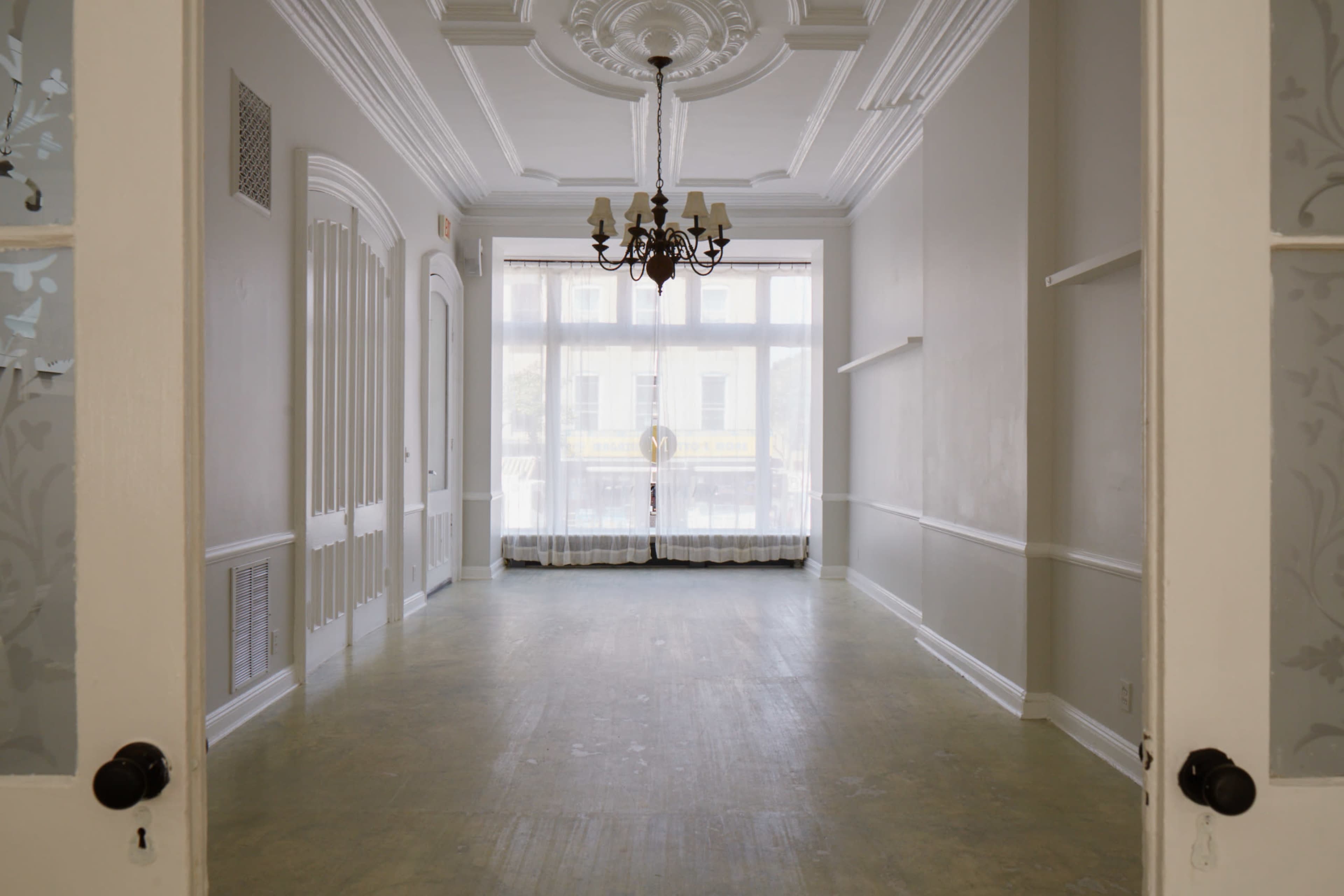 A spacious, empty room with a chandelier and large windows letting in natural light.