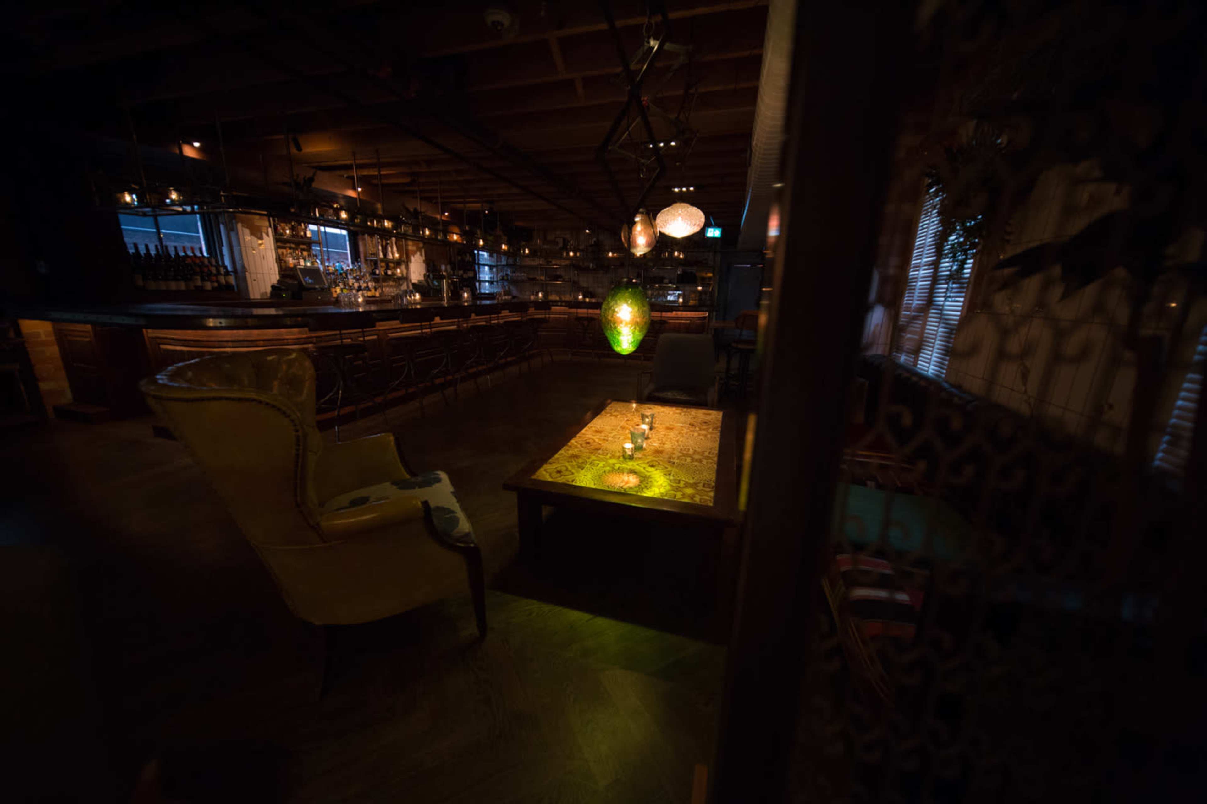 The image shows a dimly lit restaurant interior with a wooden bar, a decorative table in the foreground, and seating arranged around the space.