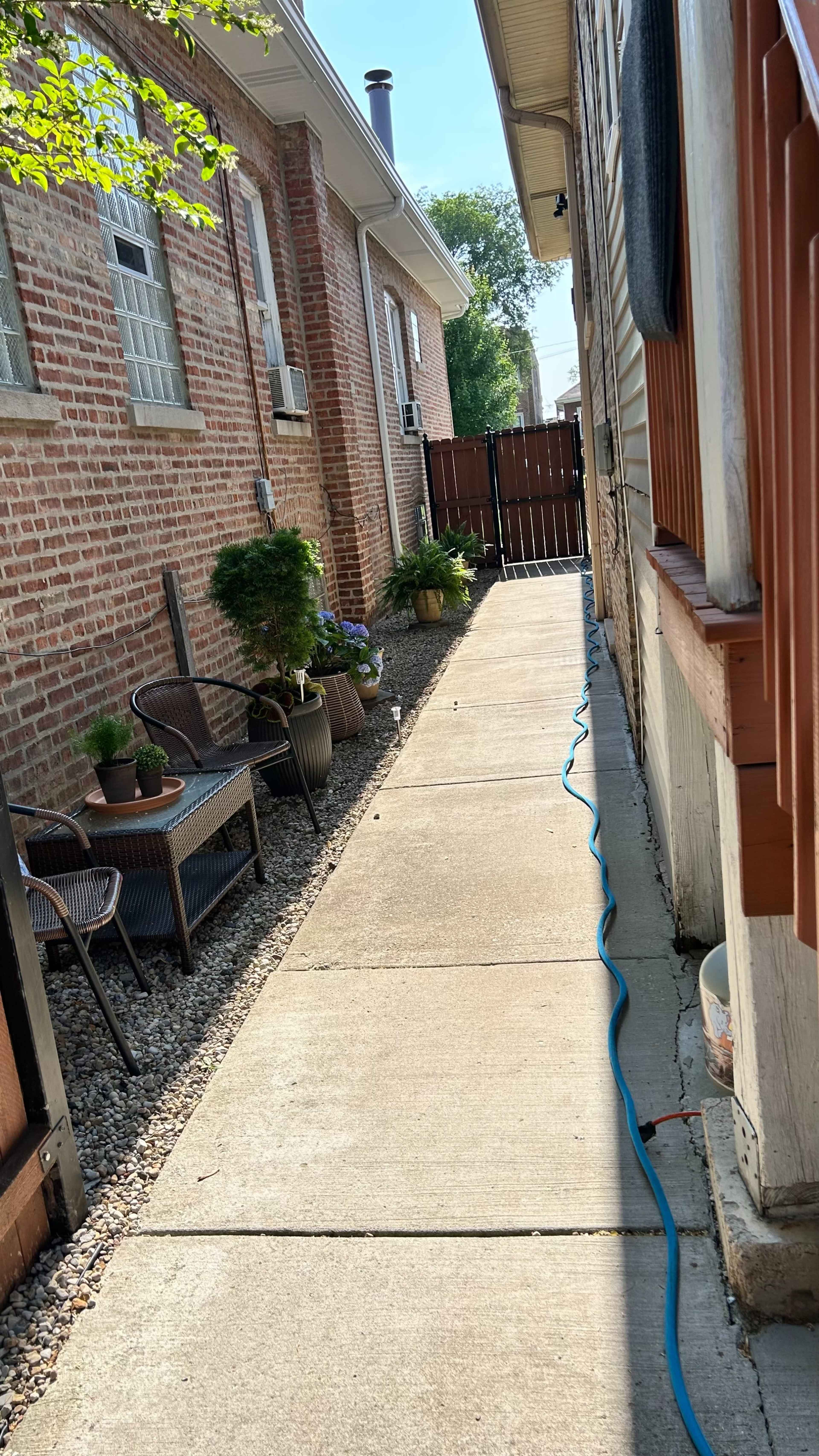 The image shows a narrow pathway between two brick buildings, lined with potted plants and leading to a fenced area at the end.