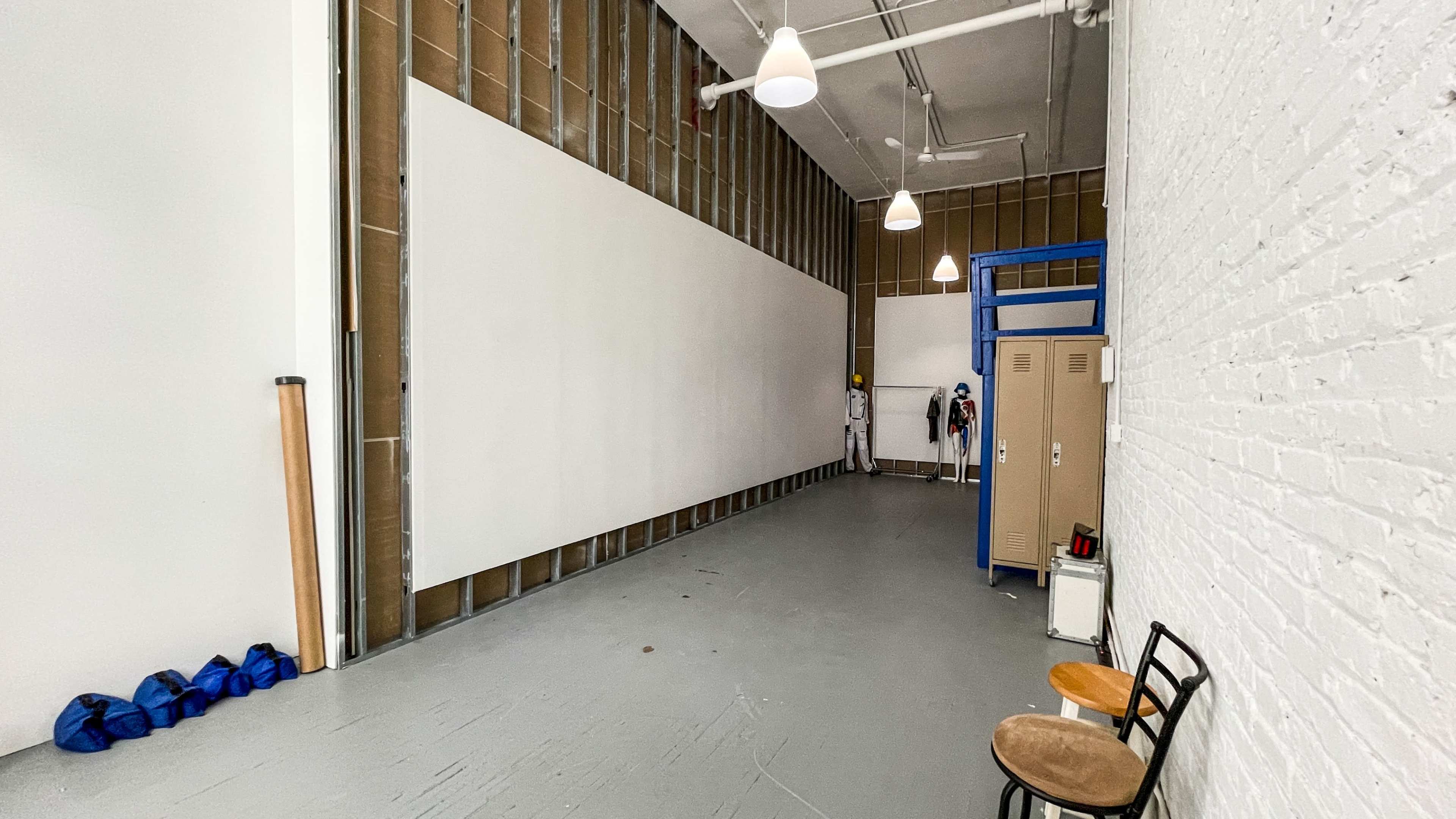 The image shows a spacious, partially finished room with exposed walls, an empty storage locker, two chairs, and various equipment along the floor.