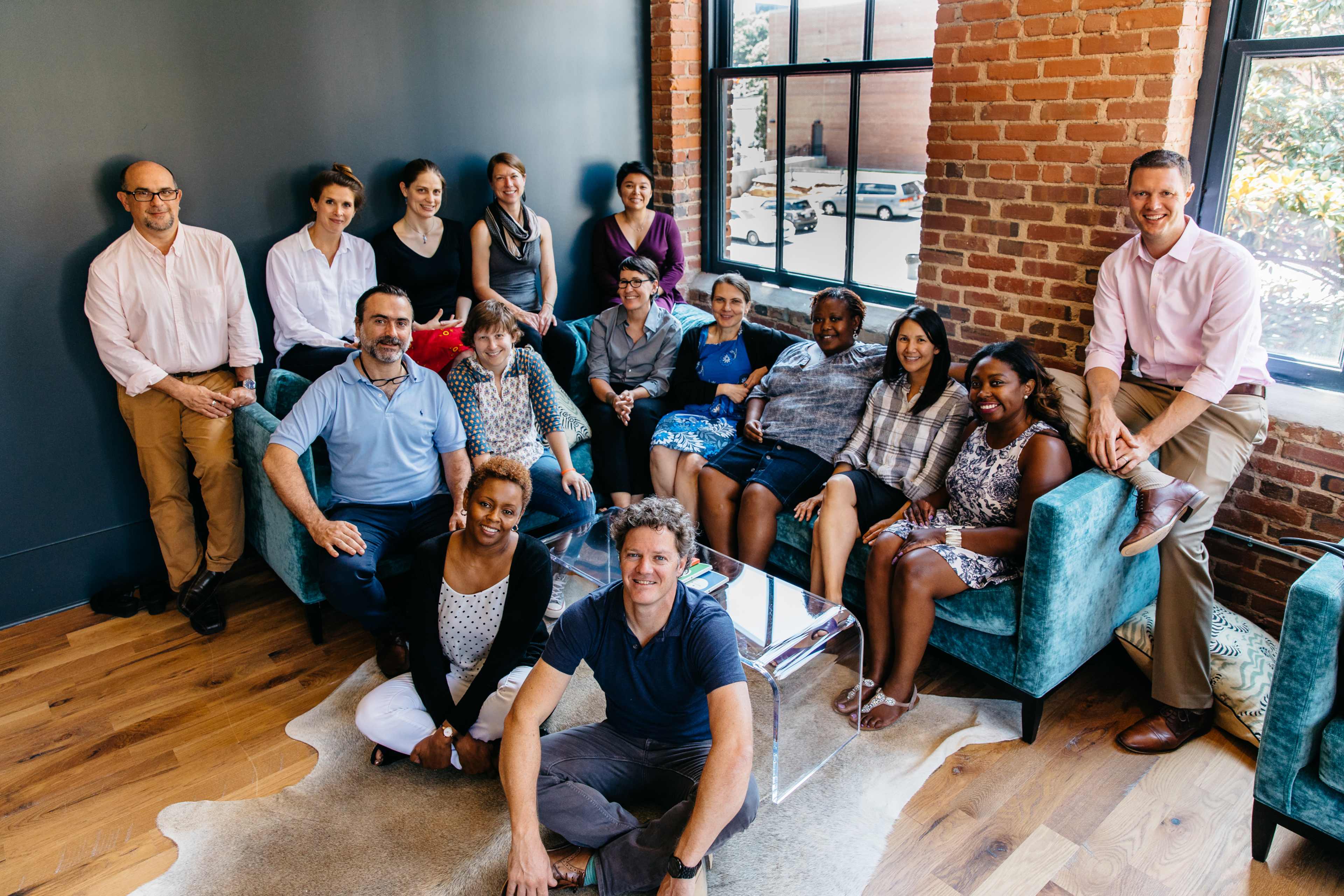 A diverse group of individuals poses for a photo in a modern, brightly lit room with brick walls and teal furniture.