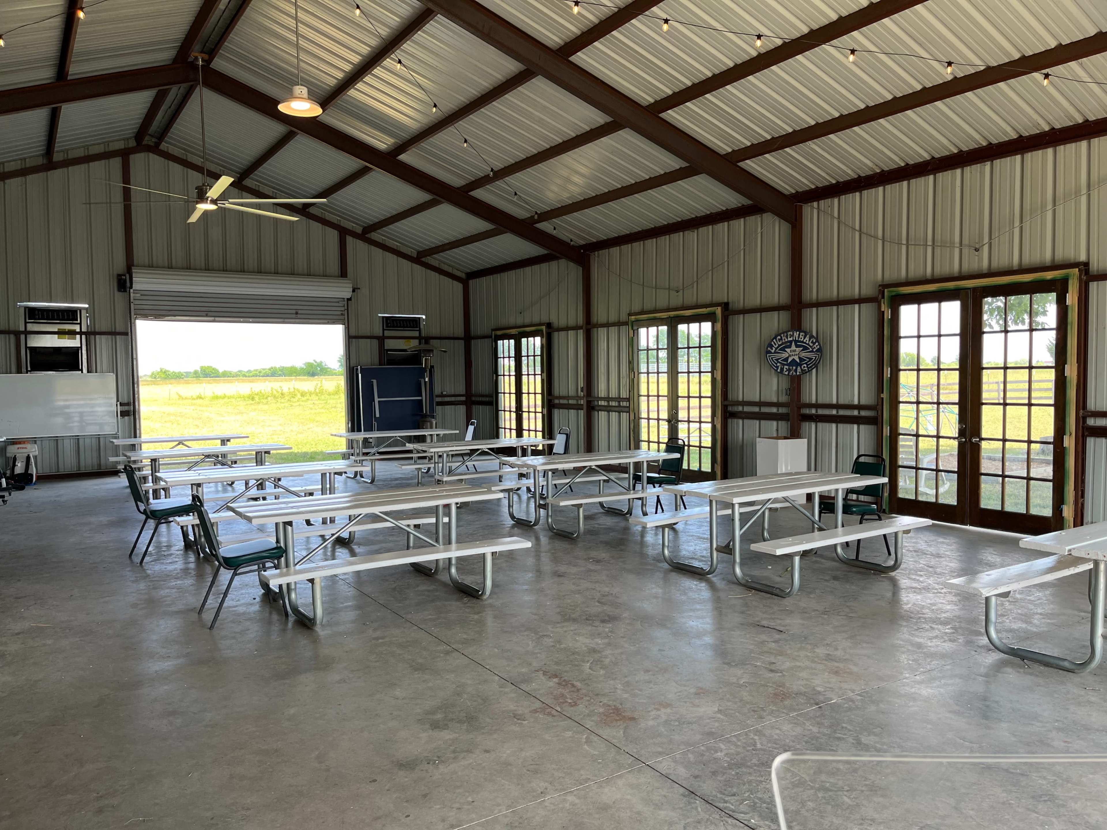 A spacious, metal-sided room with several metal folding tables and chairs arranged for a gathering, featuring large windows that allow natural light and a view of the outdoors.