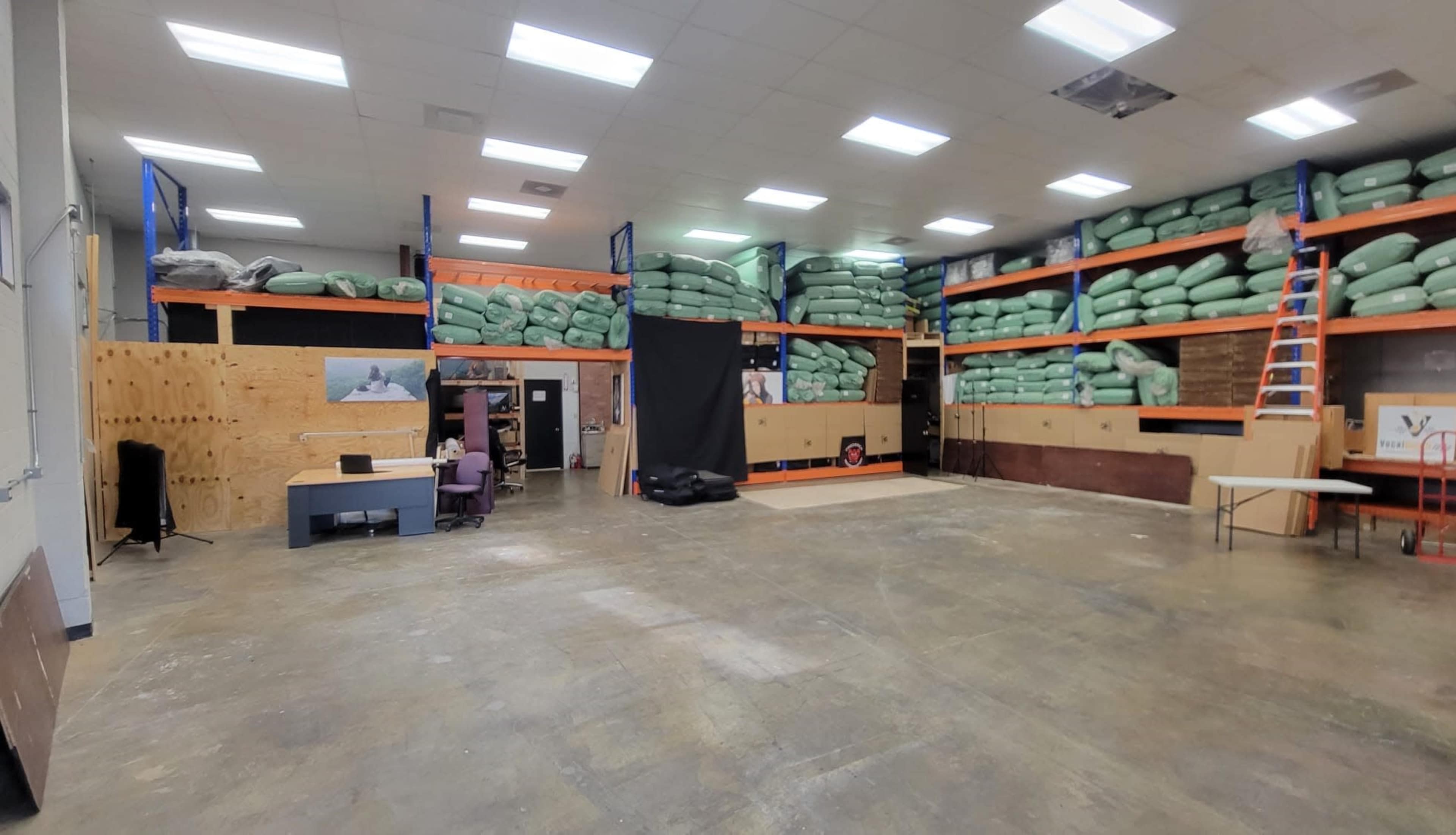 A spacious warehouse interior with stacked green bags on shelves and a workspace featuring a desk and chairs alongside a black backdrop.