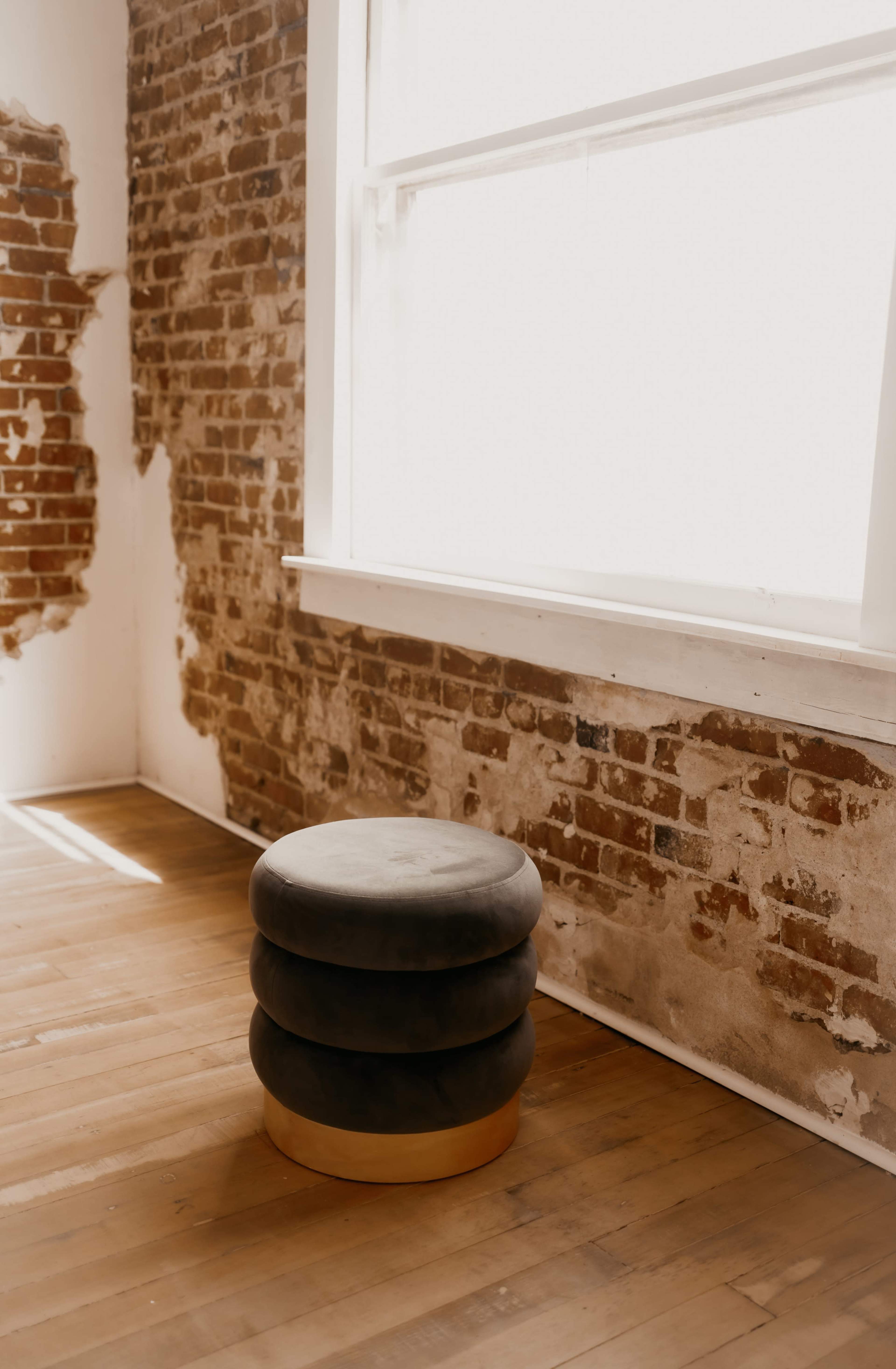 A round, stacked stool sits on a wooden floor beside a large window, with exposed brick walls in the background.