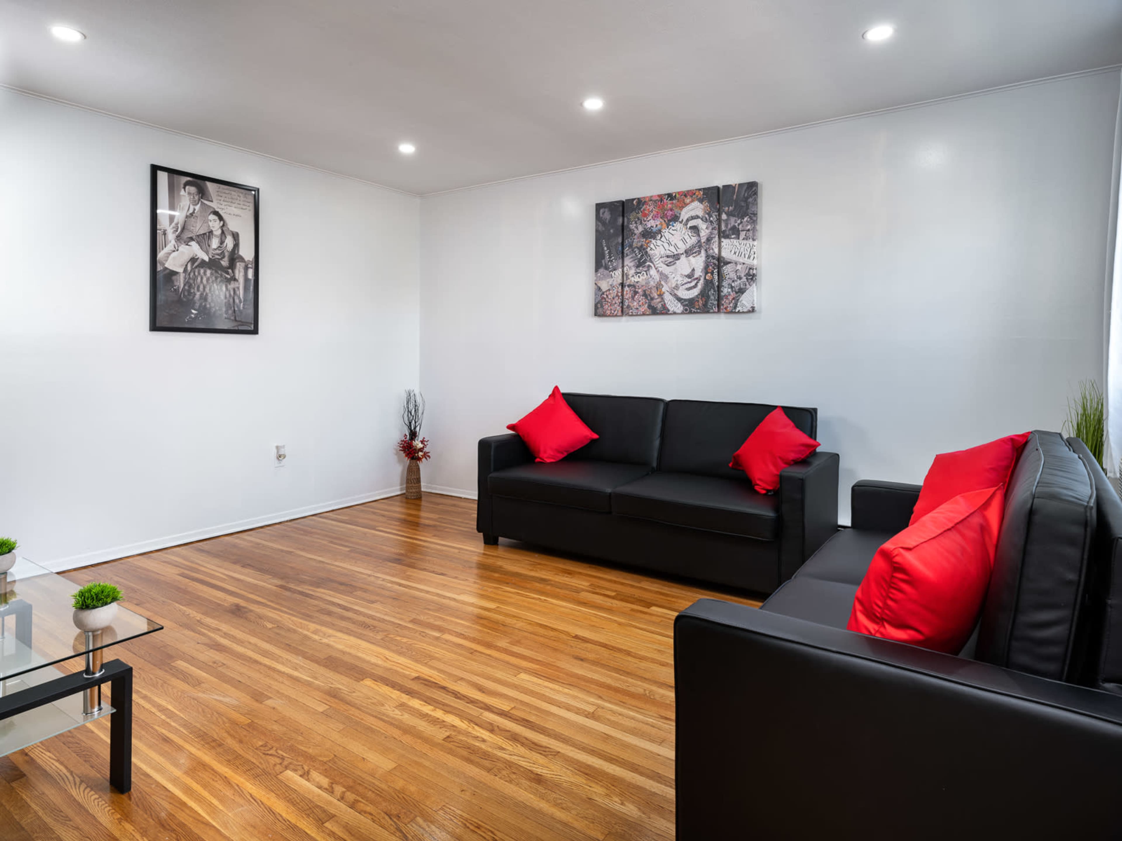 A living room with two black sofas adorned with red cushions, hardwood flooring, and wall art featuring black-and-white photographs.