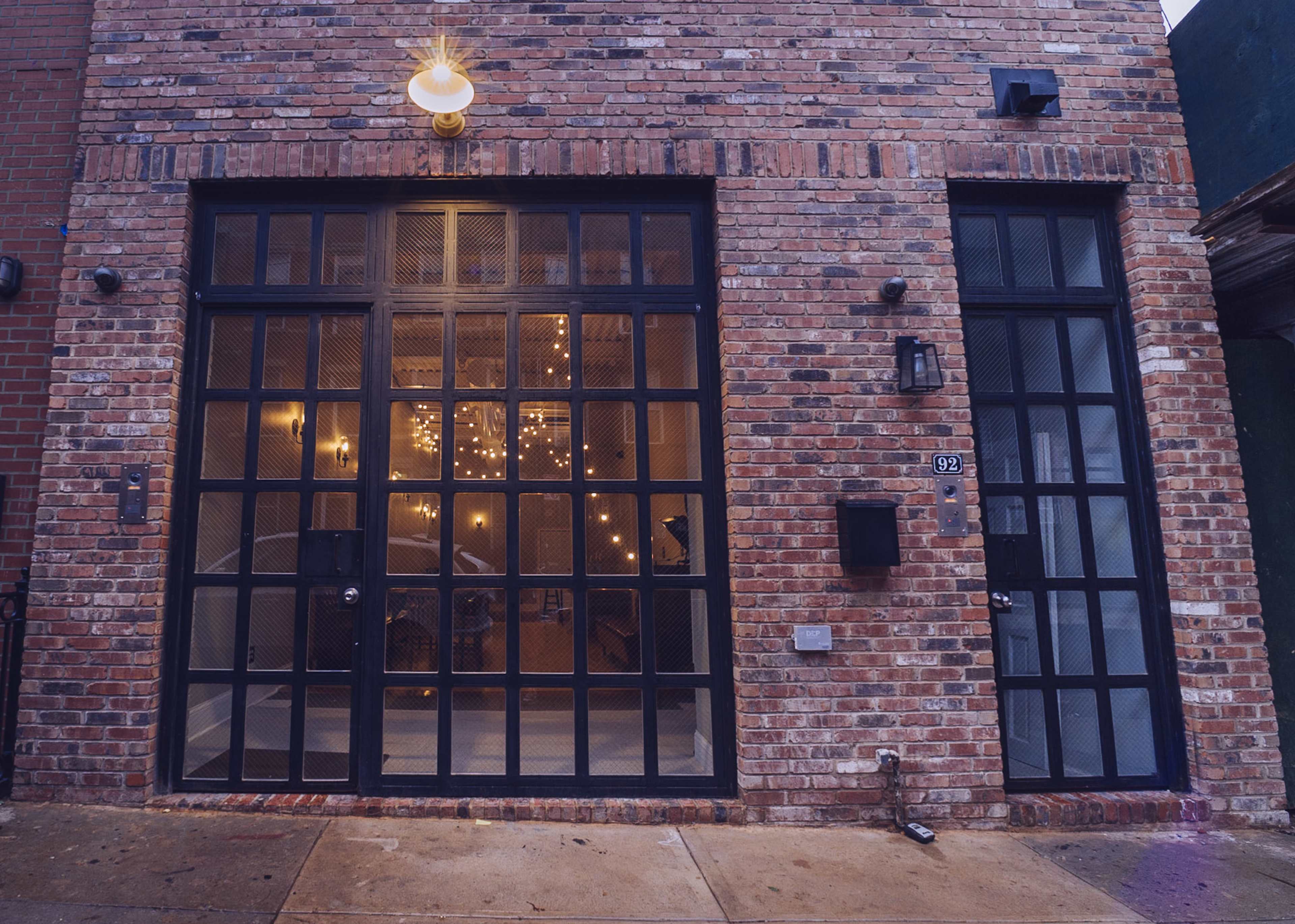 The image shows a brick building facade with large, black-framed glass doors illuminated by a wall-mounted light.