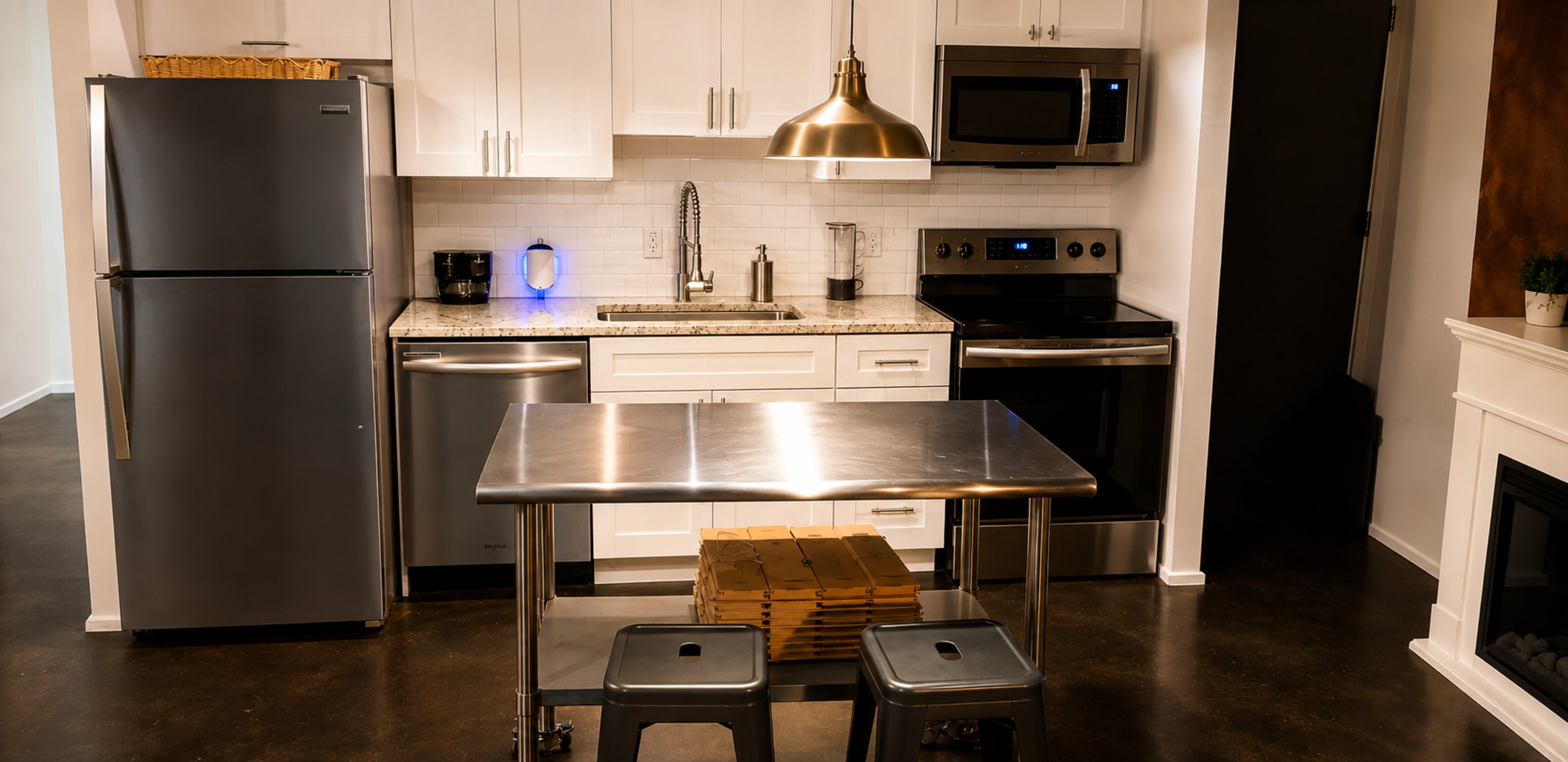 The image shows a modern kitchen with stainless steel appliances, white cabinetry, and a central stainless steel table with two metal stools.
