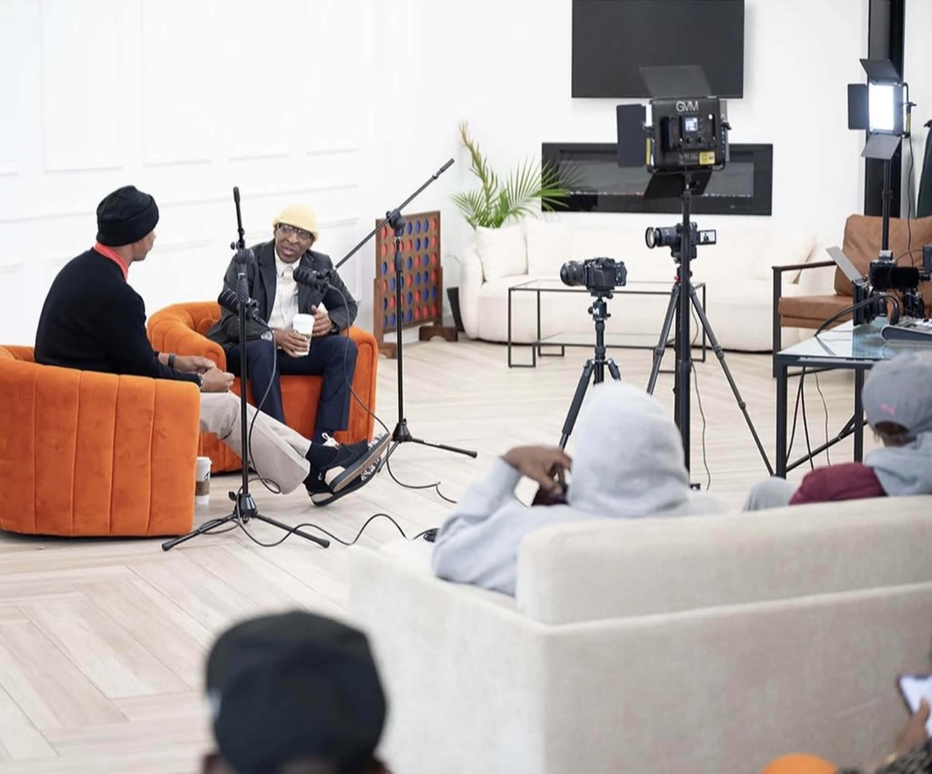 Two men are seated in an orange armchair and a black leather chair, engaged in an interview with cameras and microphones positioned around them.