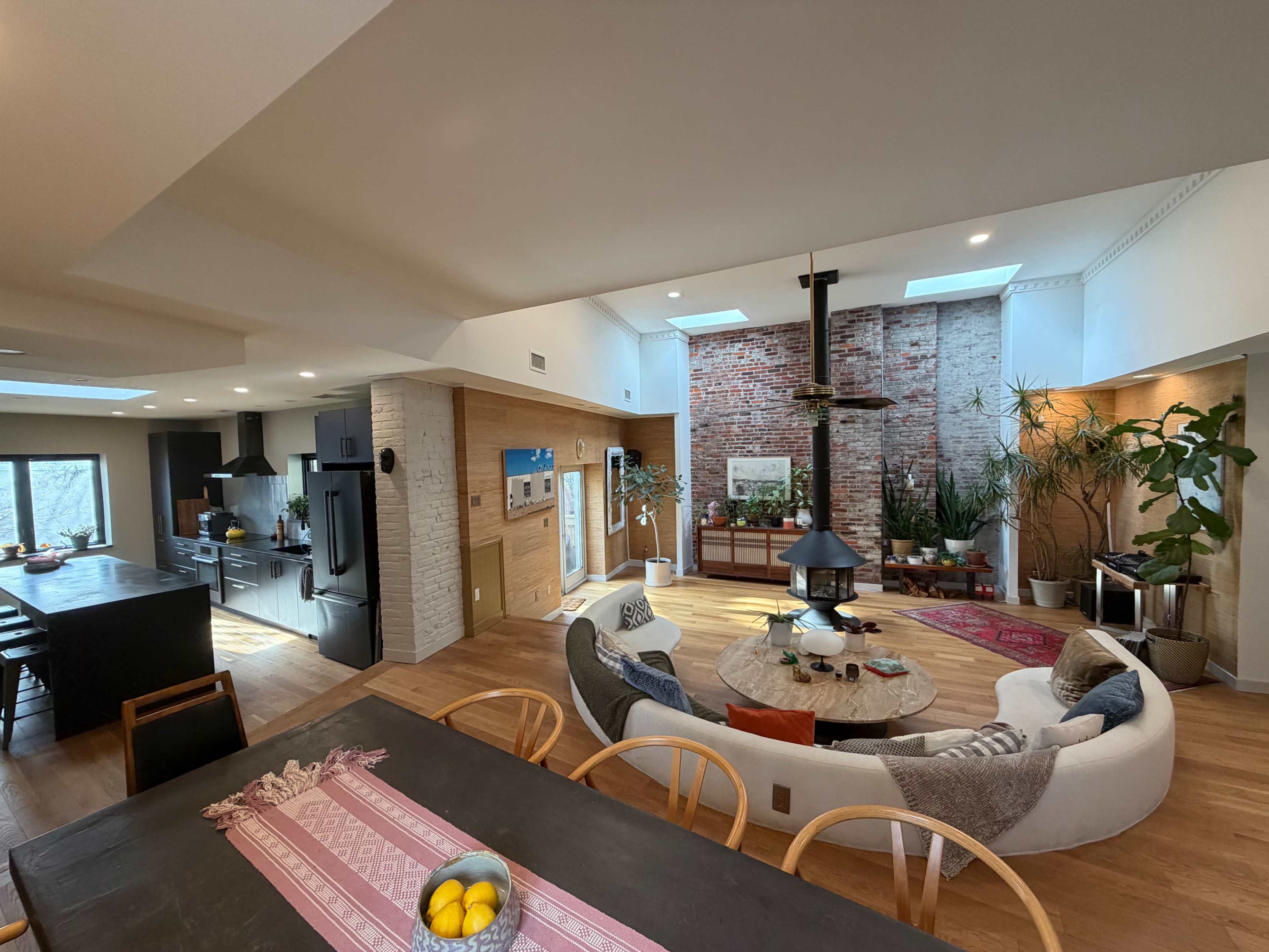 The image shows a spacious, modern living area with a circular seating arrangement, a fireplace at the center, and large windows allowing natural light to fill the room.