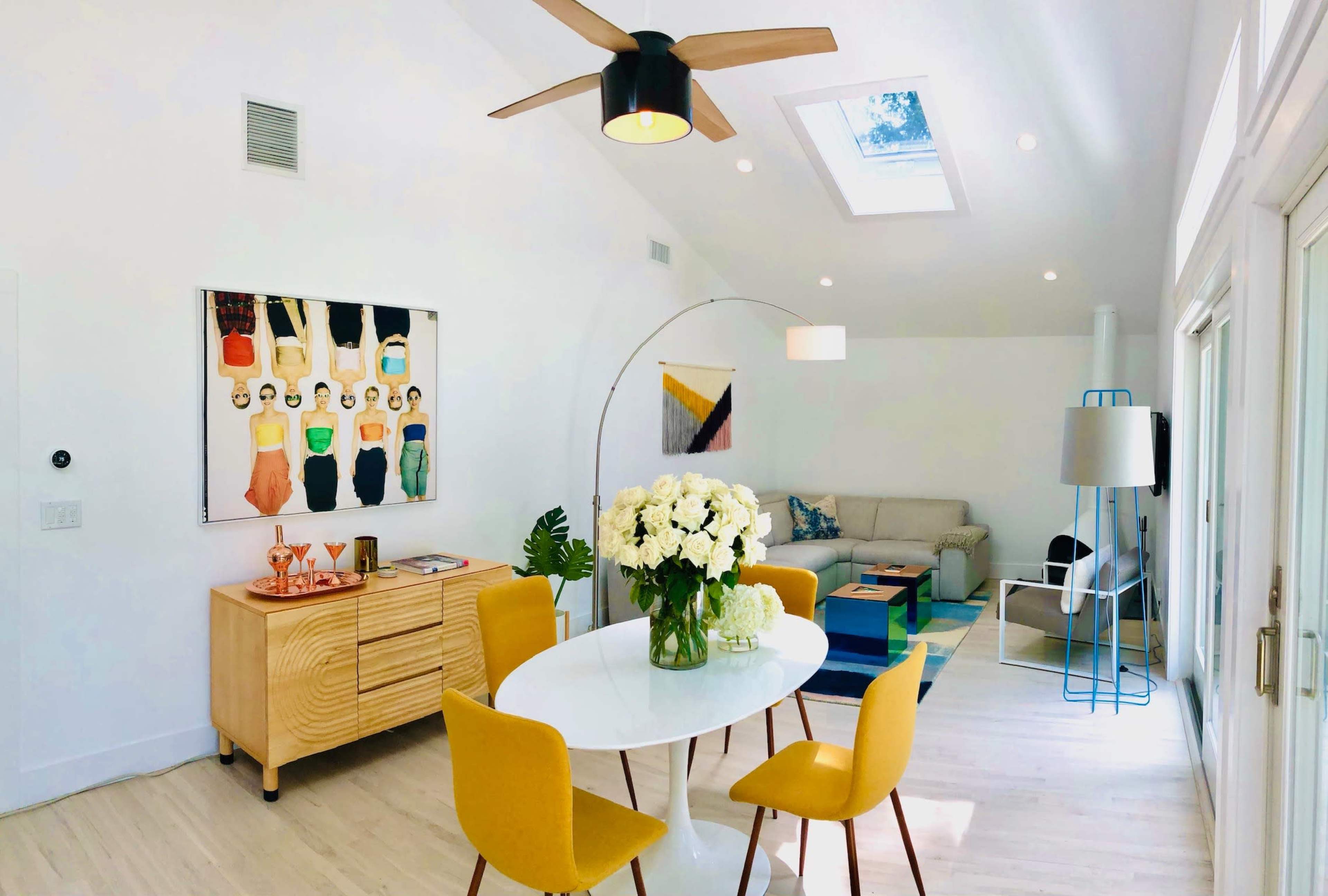 A bright room features a dining table with yellow chairs, a wooden sideboard with colorful artwork, and a cozy seating area.