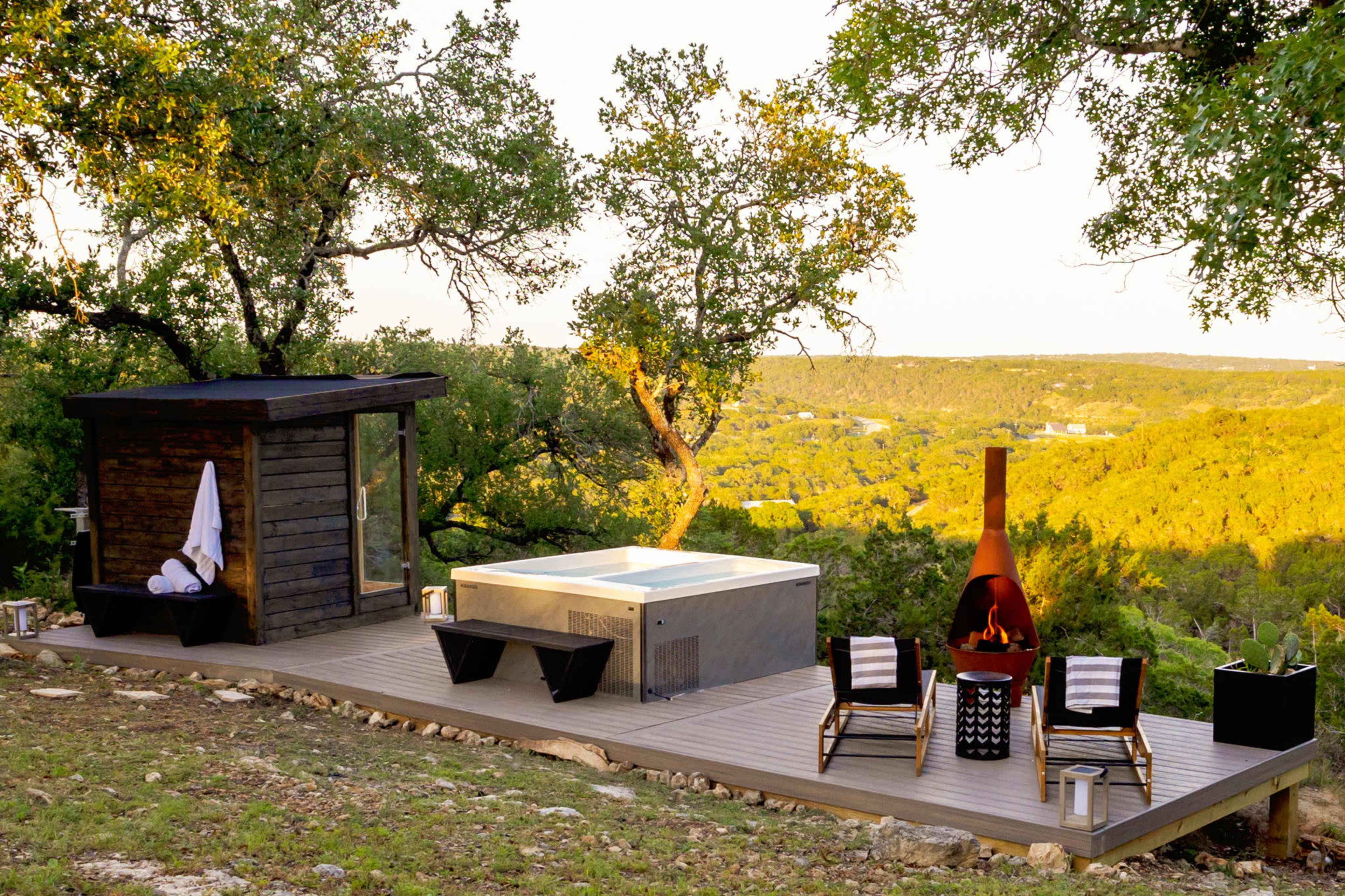 The image shows a modern outdoor deck with a hot tub, seating area, and a small wooden structure surrounded by trees and hills.