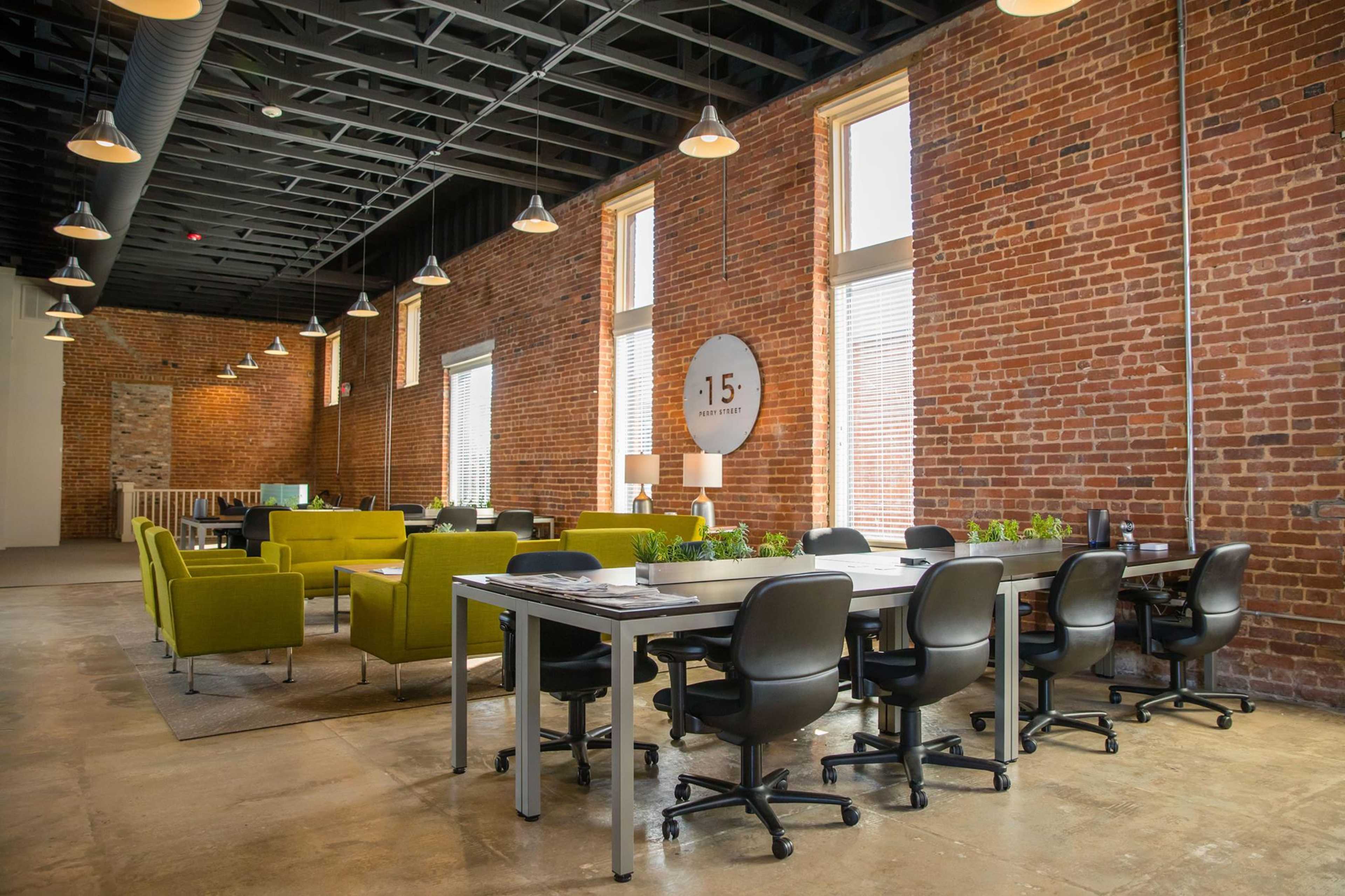 A modern office space with exposed brick walls, large windows, and a mix of green chairs and work tables.