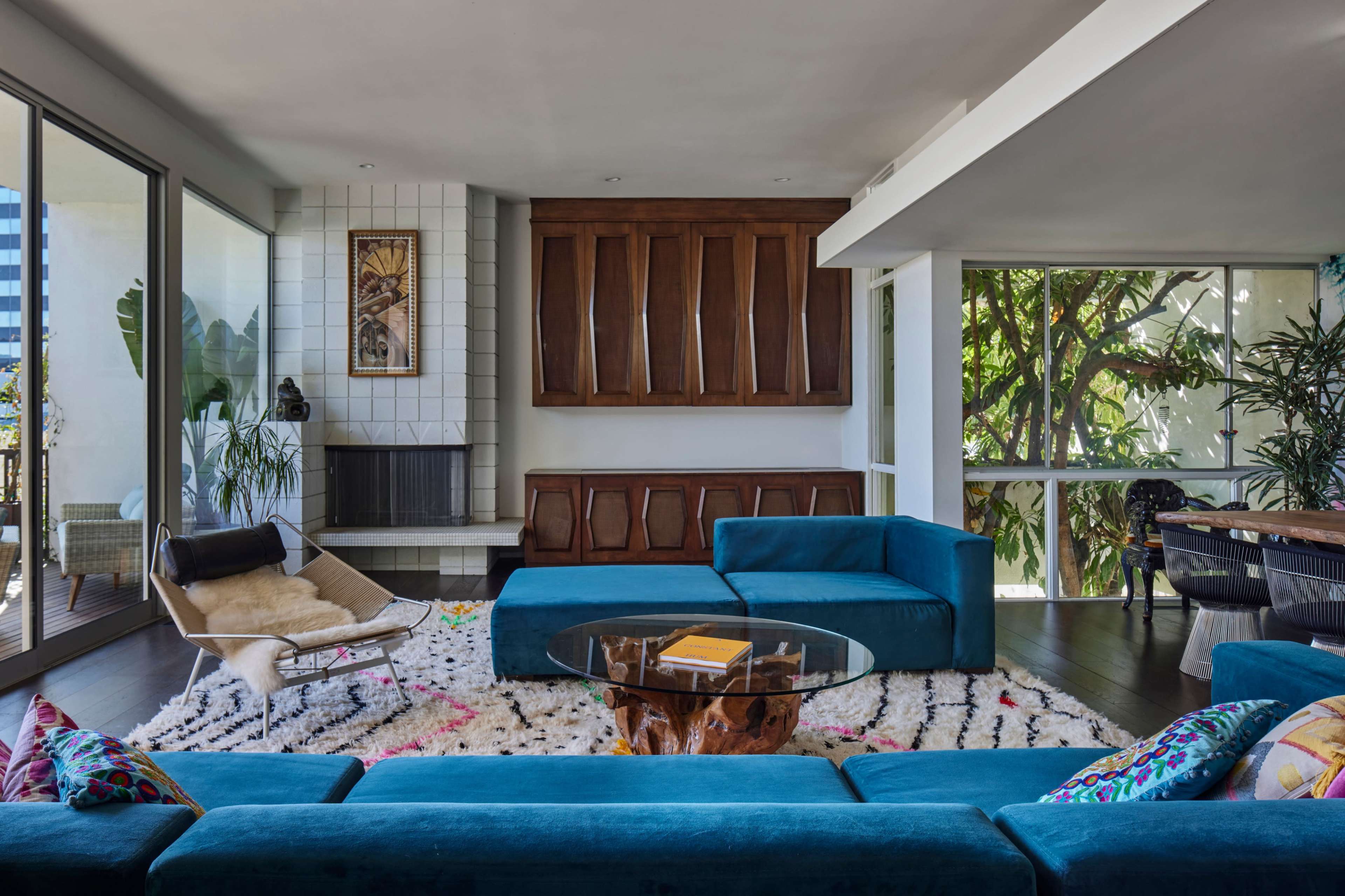 The image shows a modern living room with turquoise sofas, a round glass coffee table, a fireplace, and large windows allowing natural light.