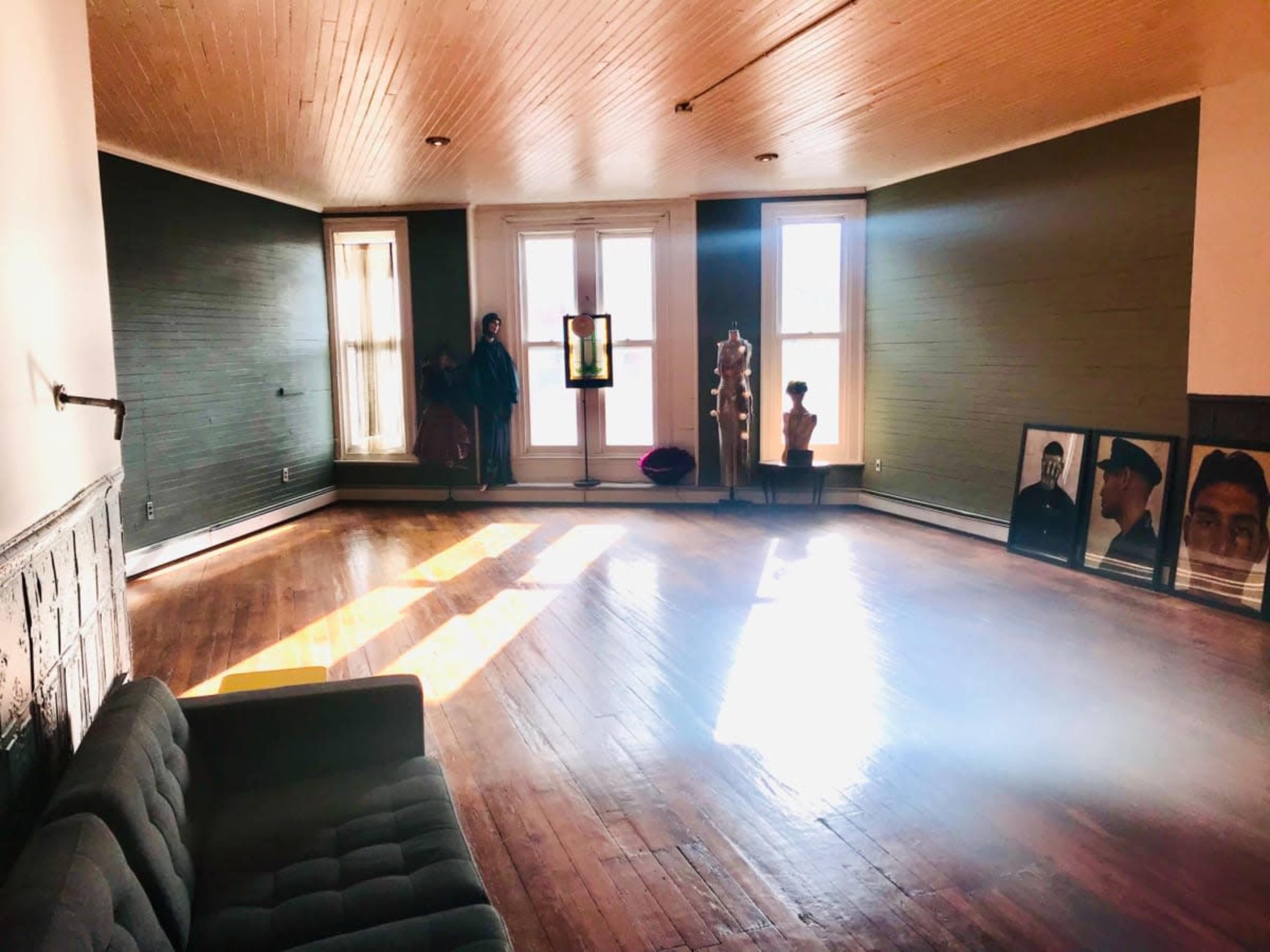 The image shows an empty room with a wooden floor, a gray couch, two large windows allowing sunlight to enter, and various decorative elements, including a mannequin and framed photographs.