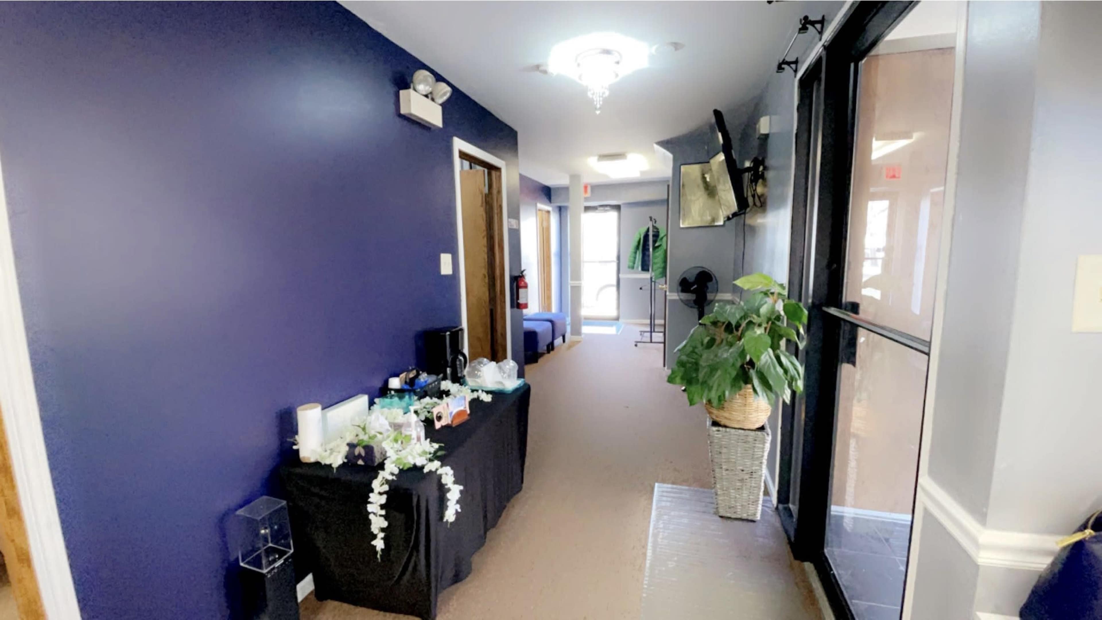 A corridor with a dark blue wall, a table covered with various items, and a doorway leading to an outdoor area.