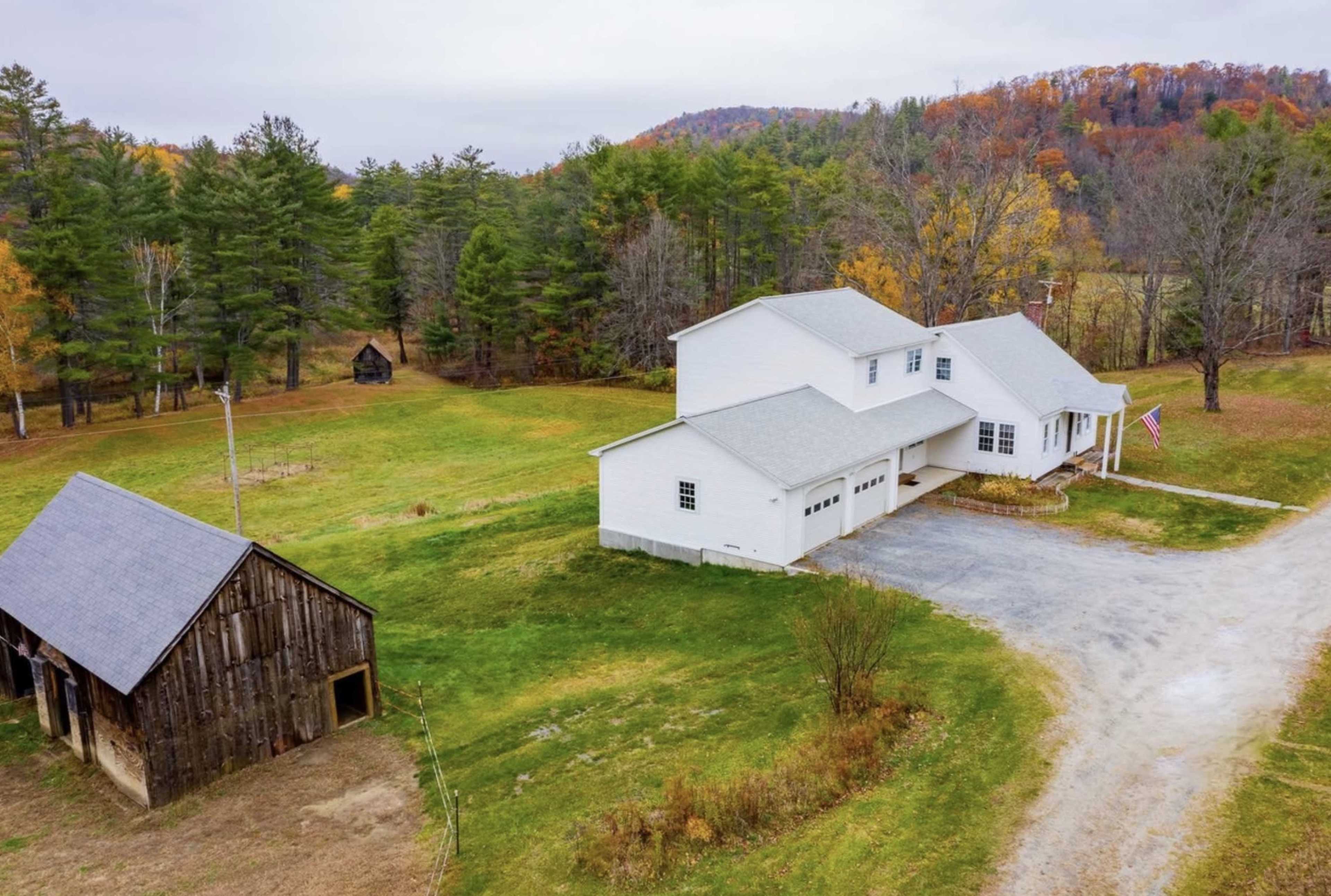 A white two-story house with a gray roof sits beside a weathered wooden barn in a rural landscape surrounded by trees and autumn foliage.
