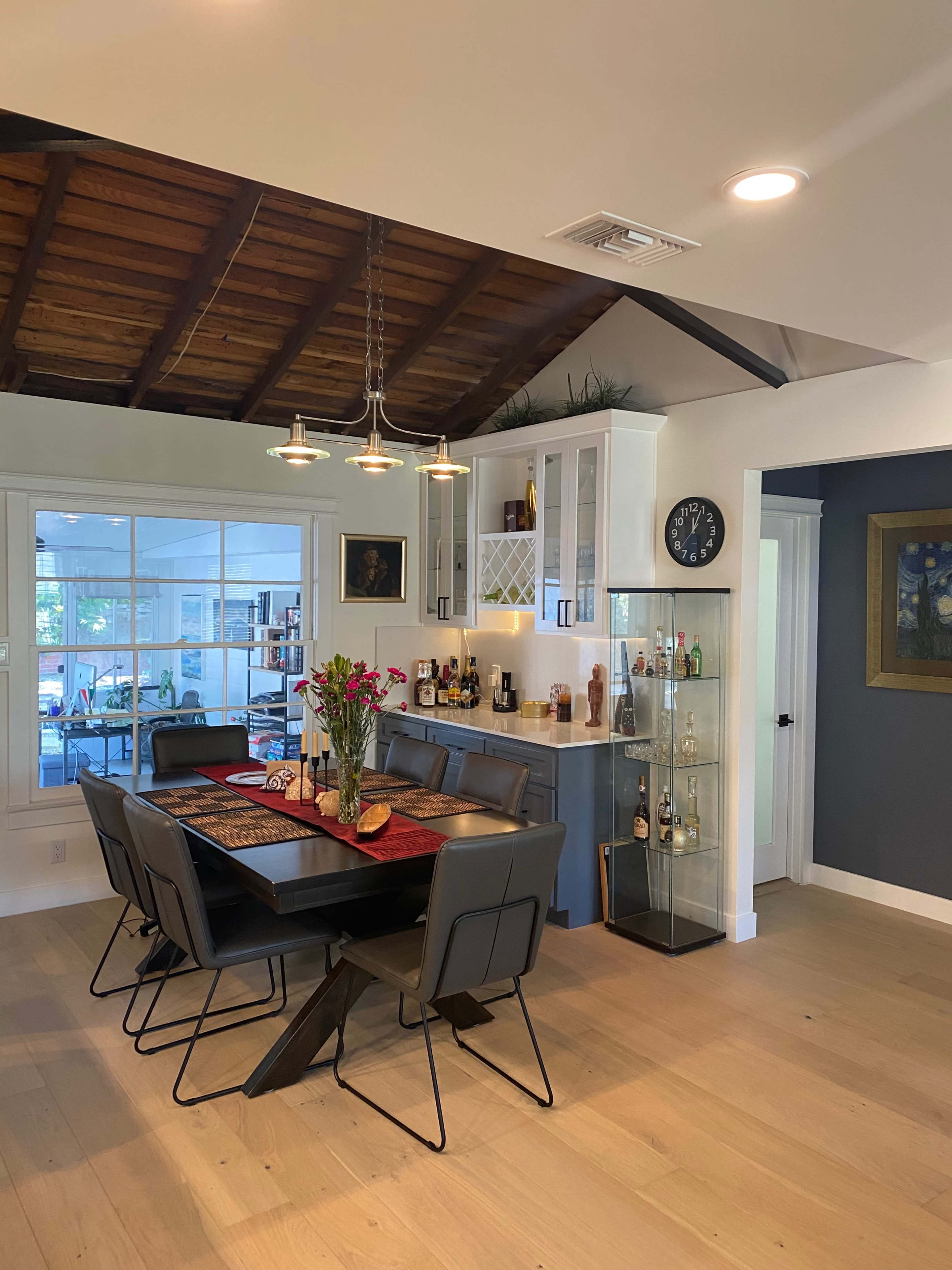 A dining area features a wooden table with black chairs, surrounded by a kitchen with modern cabinetry and a display cabinet for bottles.