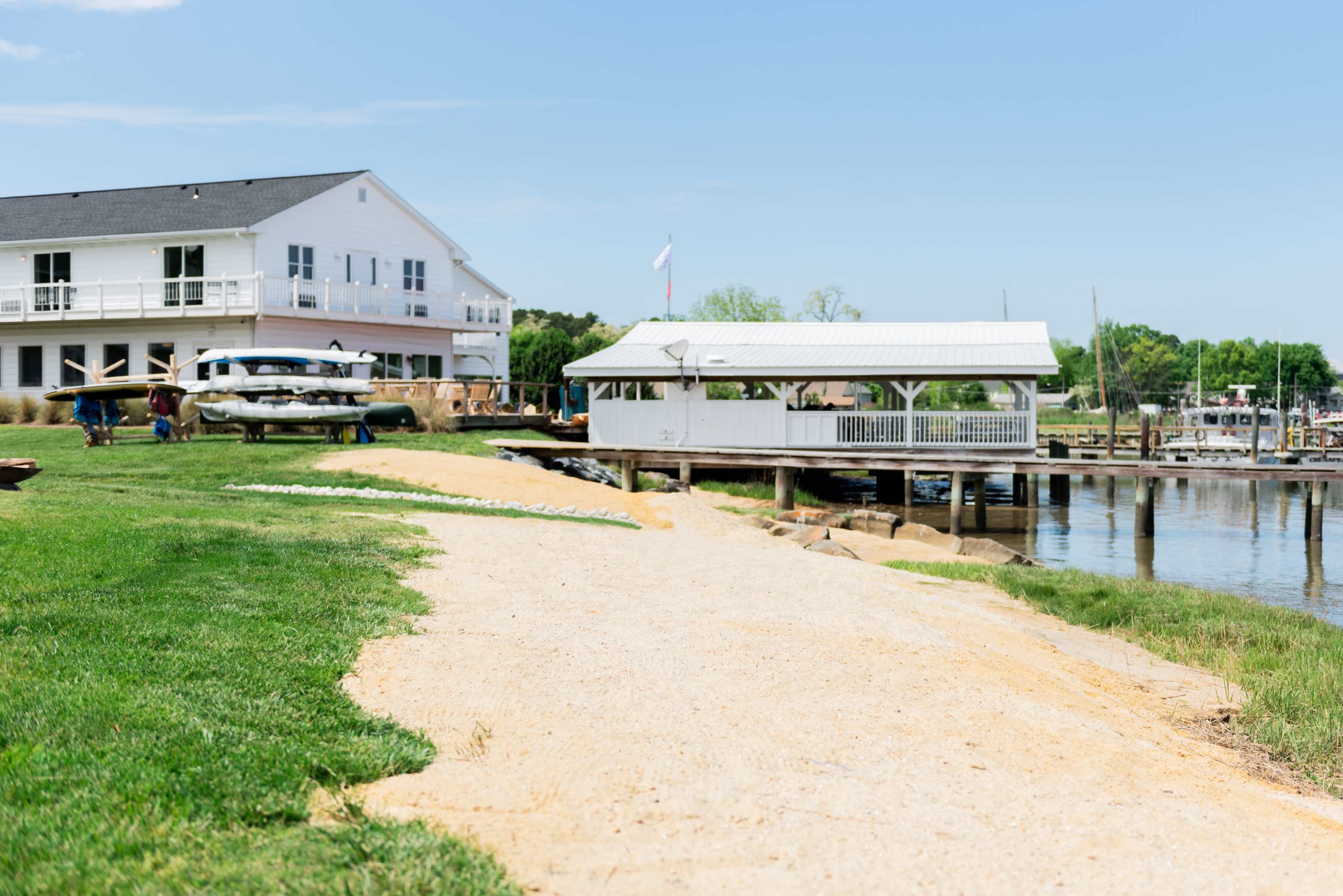 A sandy pathway leads to a dockside pavilion beside a body of water, with a white building and boats in the background.