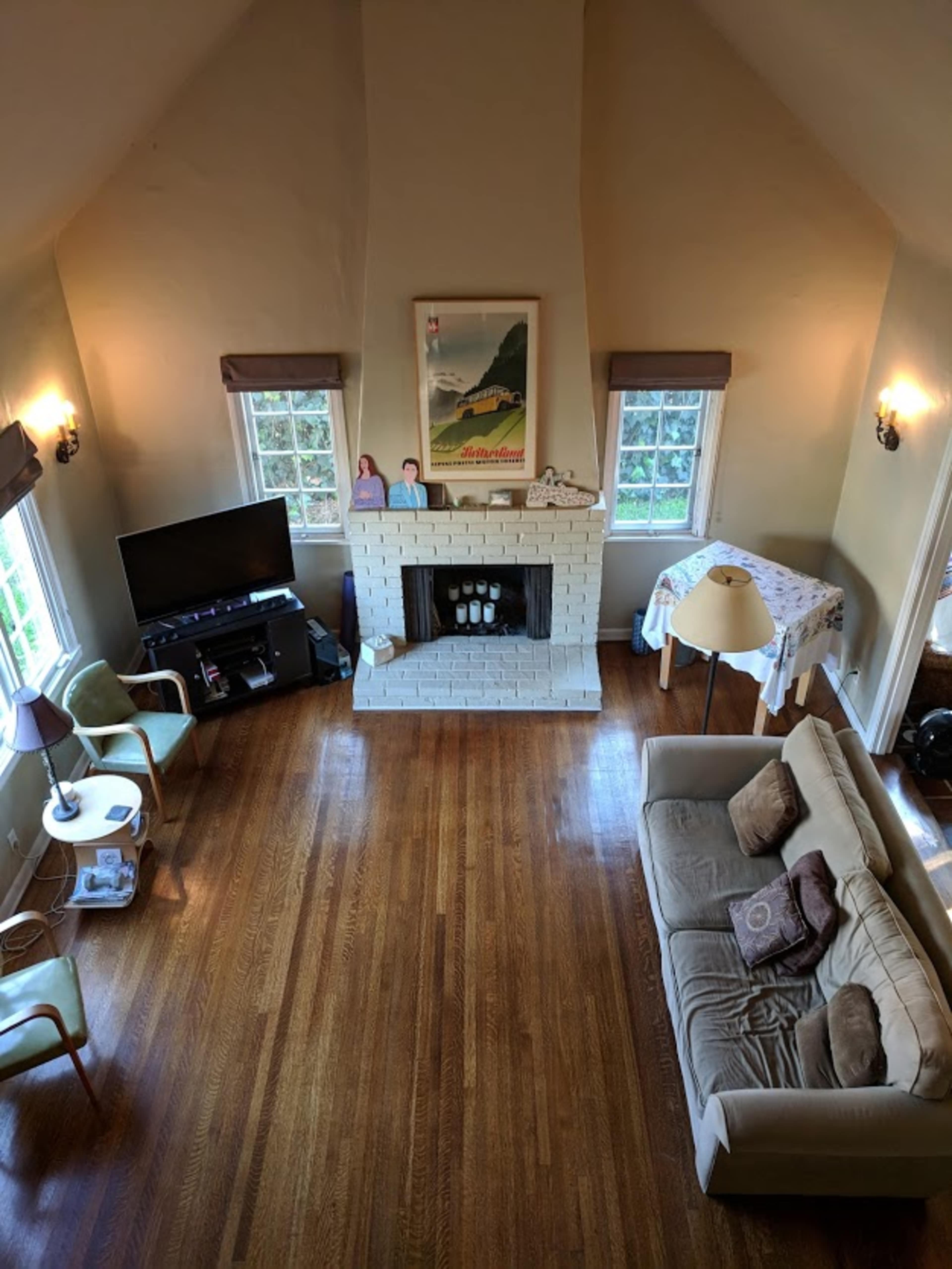 An interior of a living room with high ceilings, a white brick fireplace, two windows, a television, and a beige sofa.