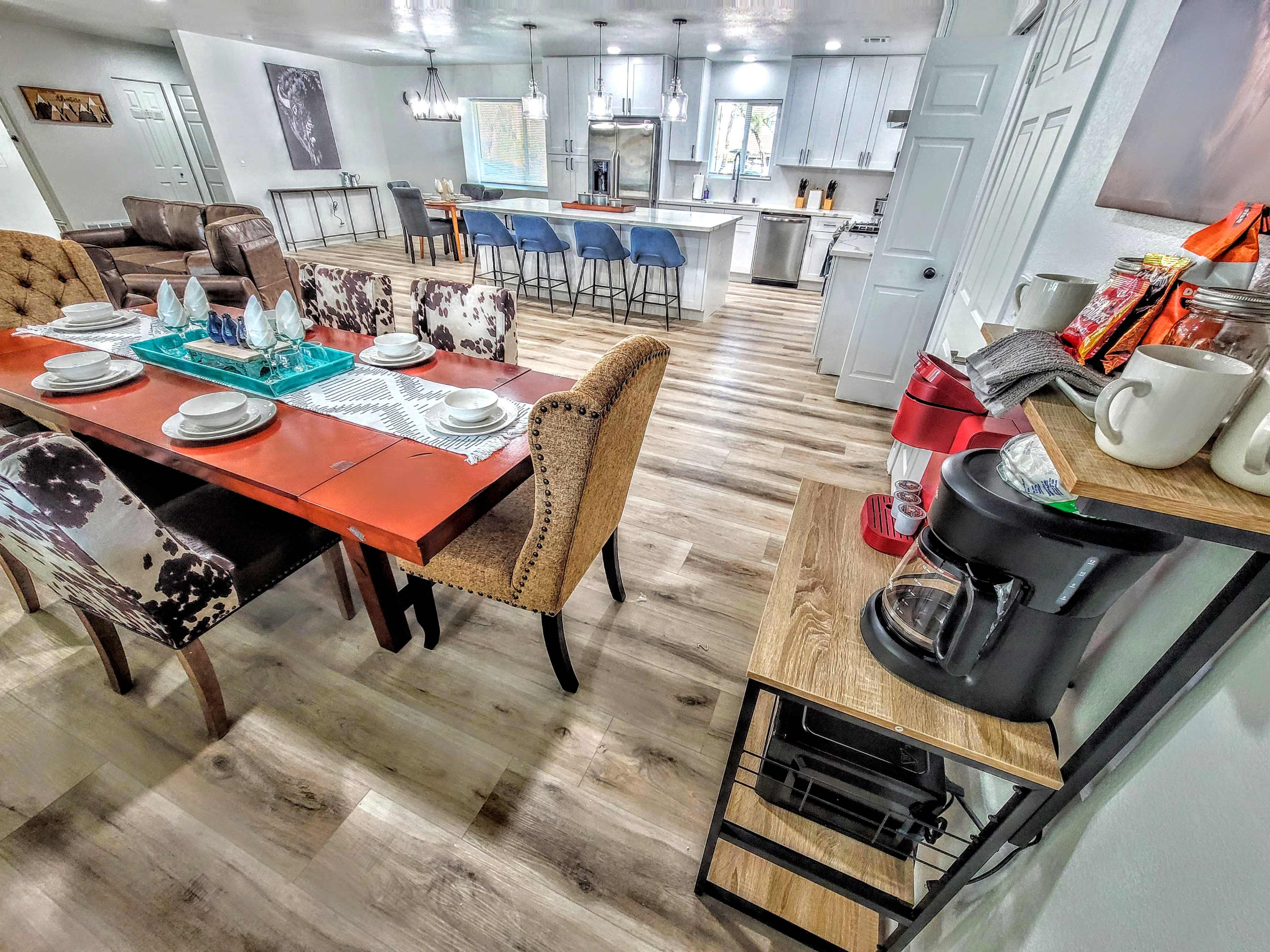 The image shows a modern kitchen and dining area featuring a red dining table set for four, a coffee station with a coffee maker, and bar stools at an island in the kitchen.