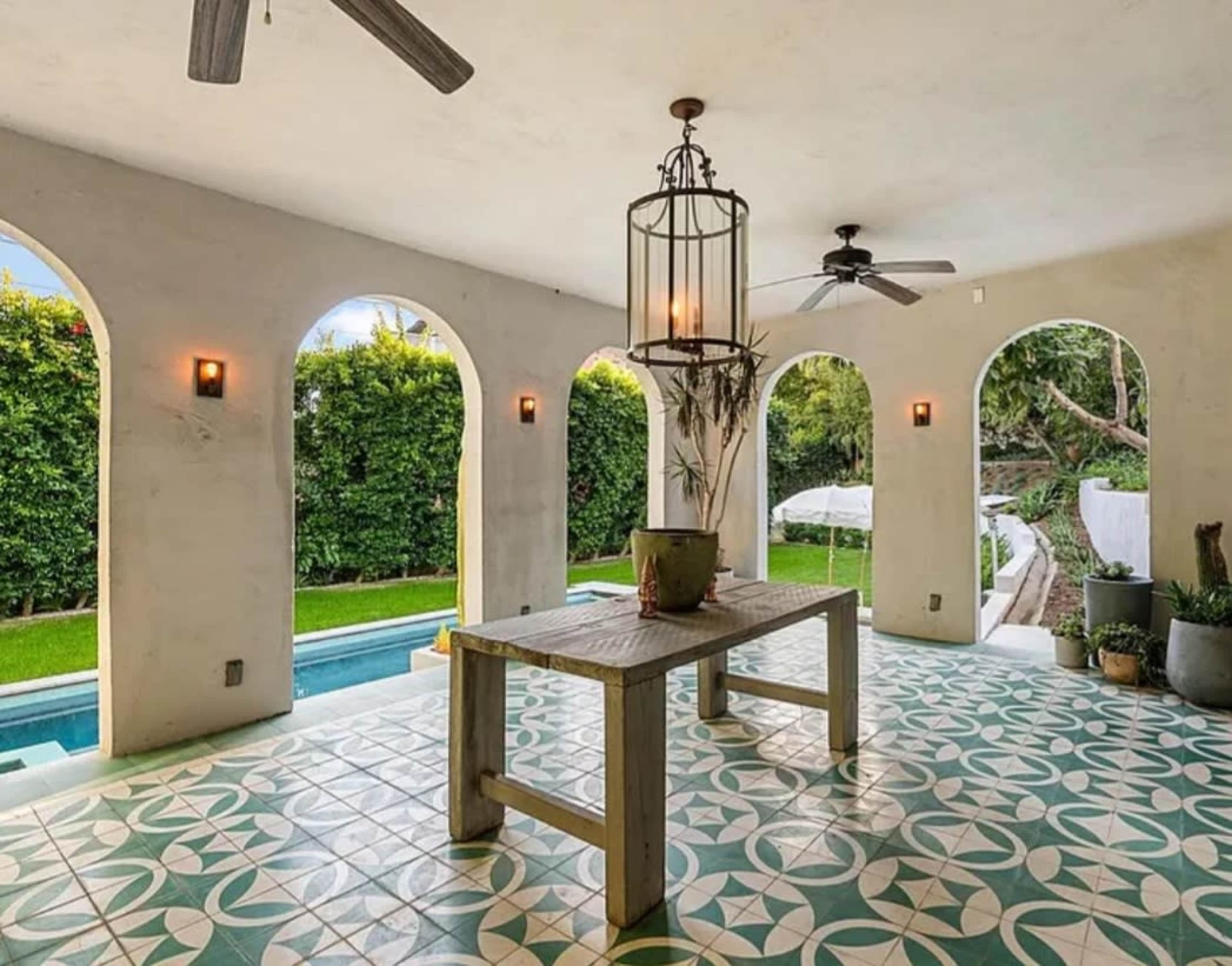 The image shows a spacious indoor area with arched windows overlooking a garden, featuring a wooden table and decorative tile flooring.