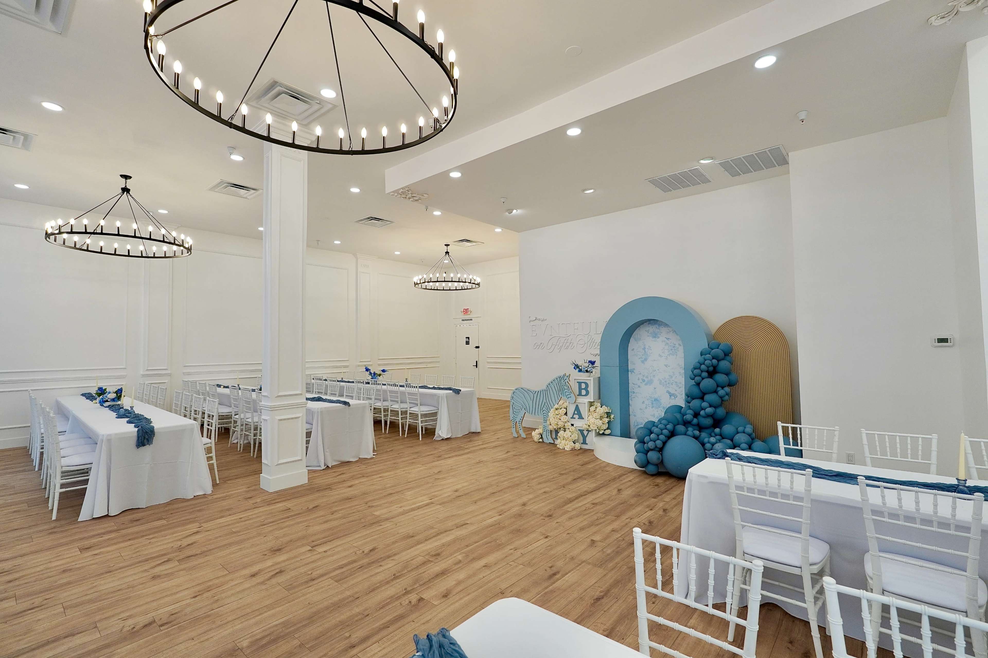 The image shows a spacious banquet room decorated with blue accents, featuring long white tables and a decorative backdrop with balloons and a themed display.