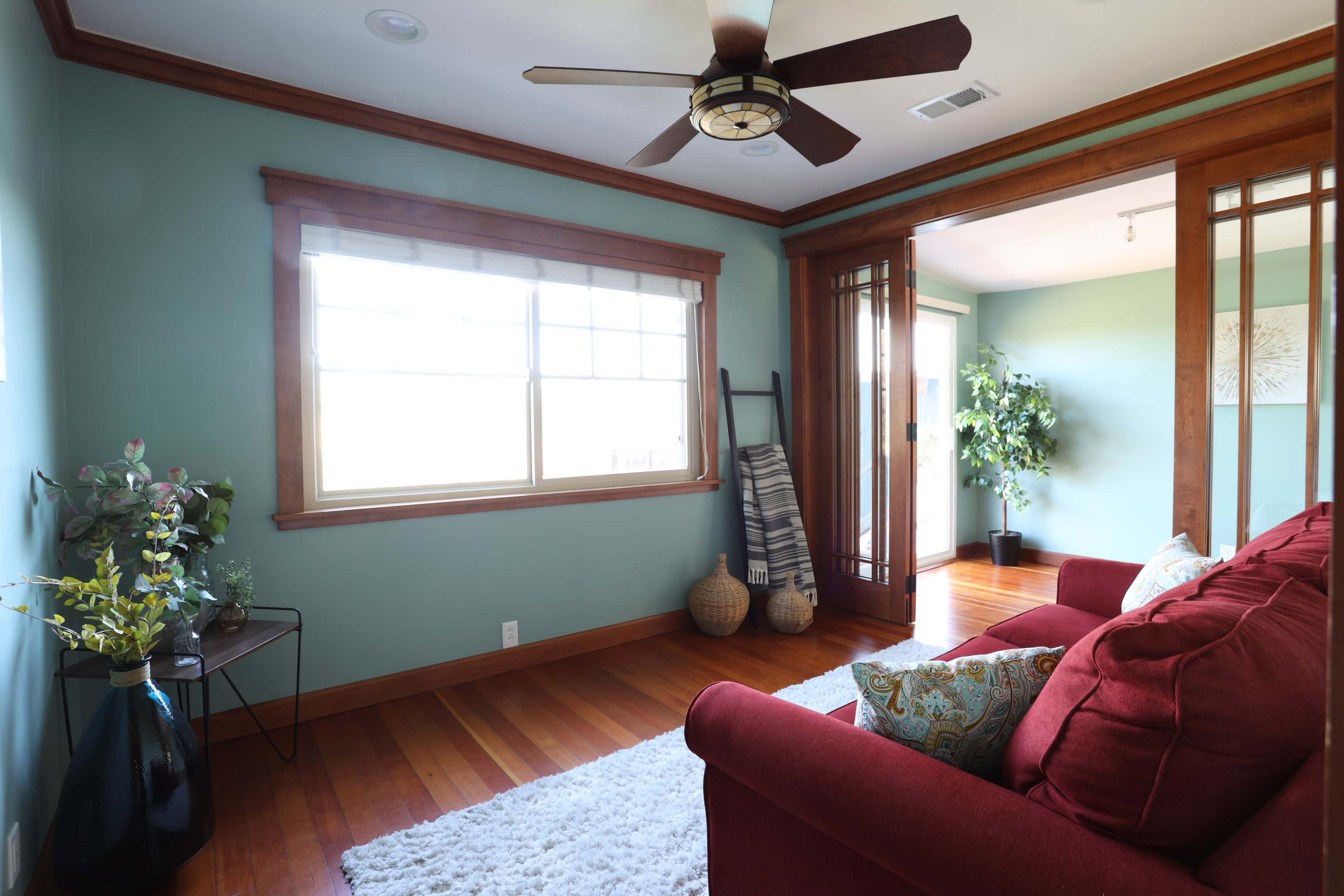 A cozy living room features a red couch, a wooden ceiling fan, and large windows with natural light.
