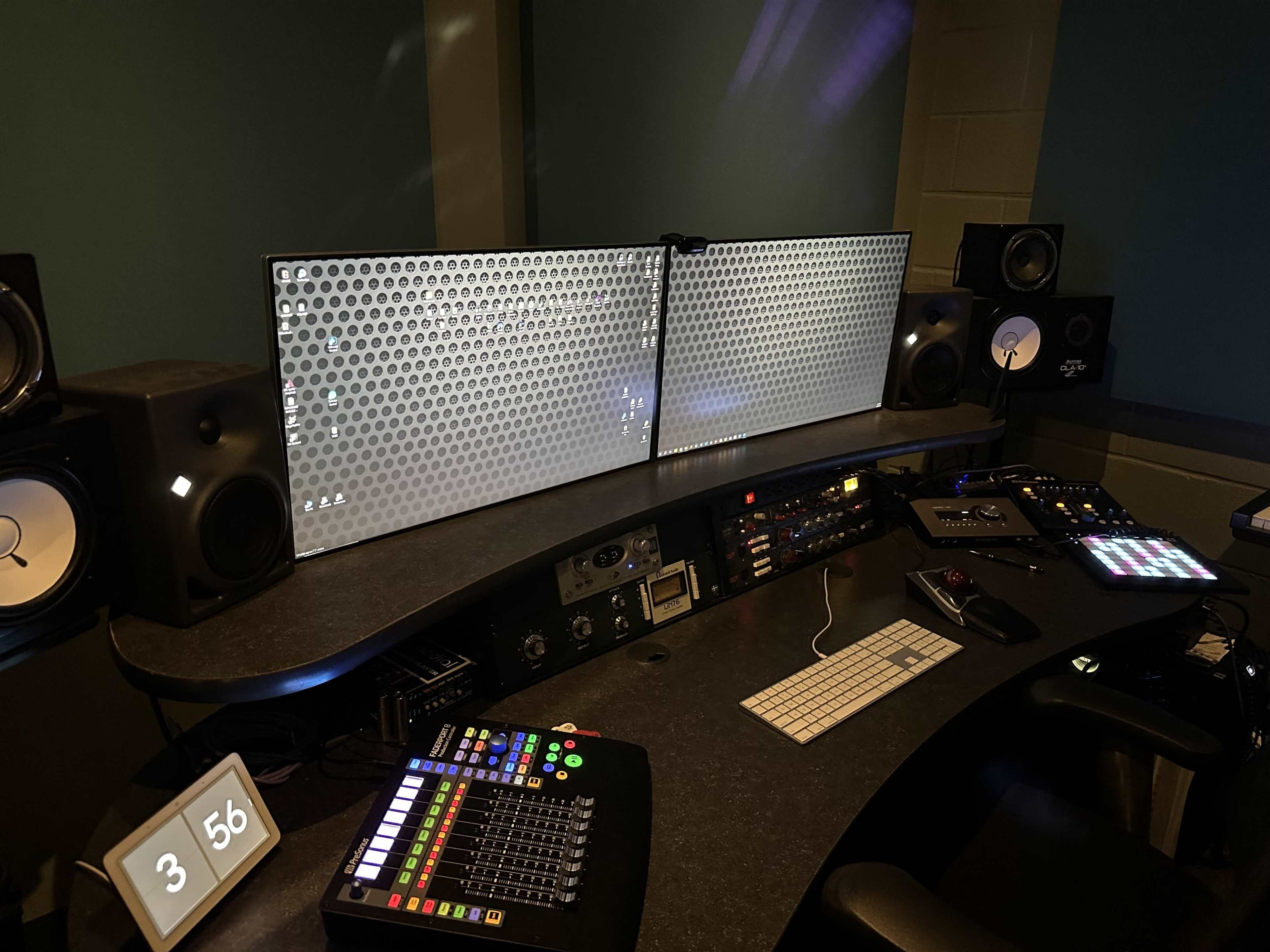 A music production studio featuring dual monitors, a mixing console, various audio equipment, and speakers arranged on a U-shaped desk.