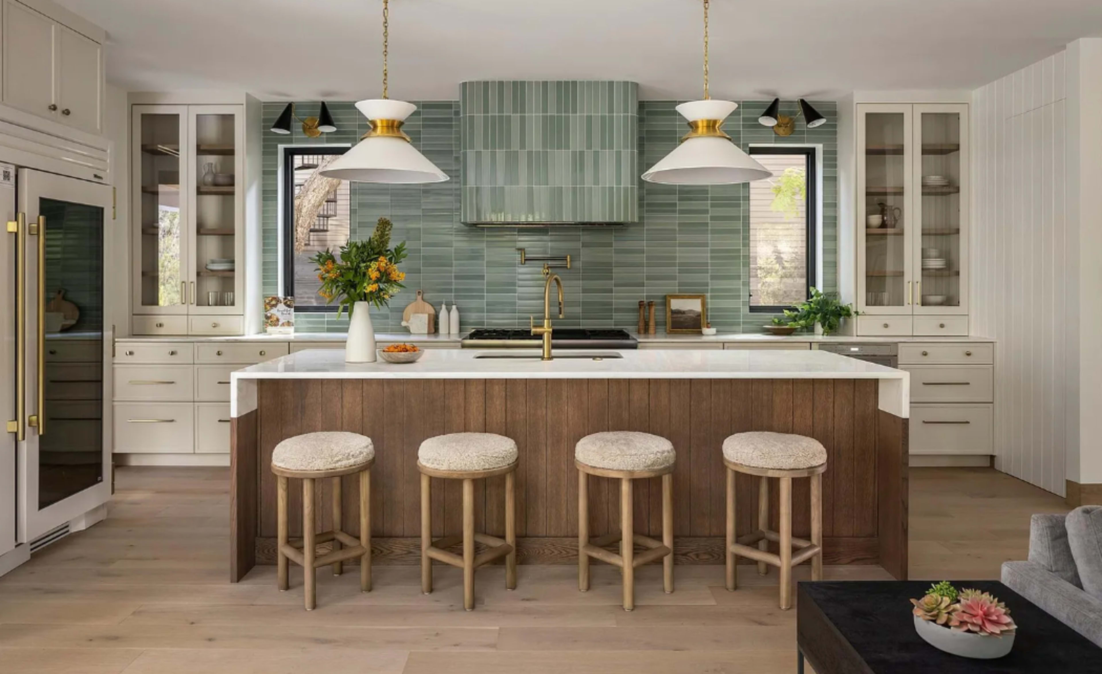 The image shows a modern kitchen featuring a large island with wooden cabinetry, four stools, and a backsplash of green tiles.