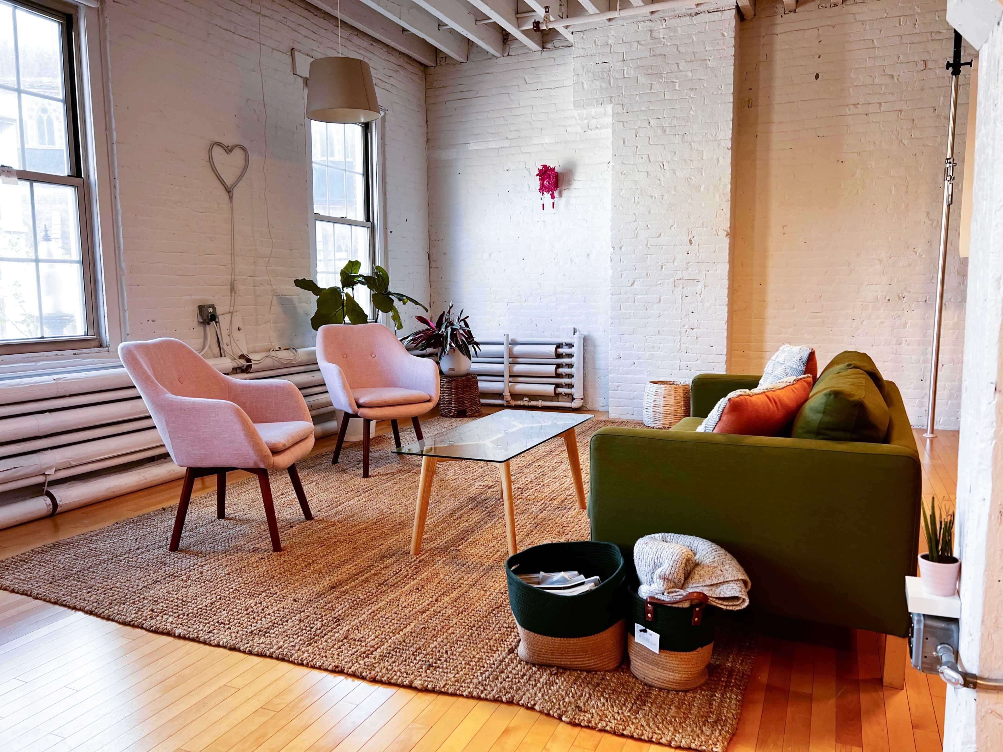 A bright, minimalist living room with two light pink chairs, a green sofa, a glass coffee table, and a woven rug.