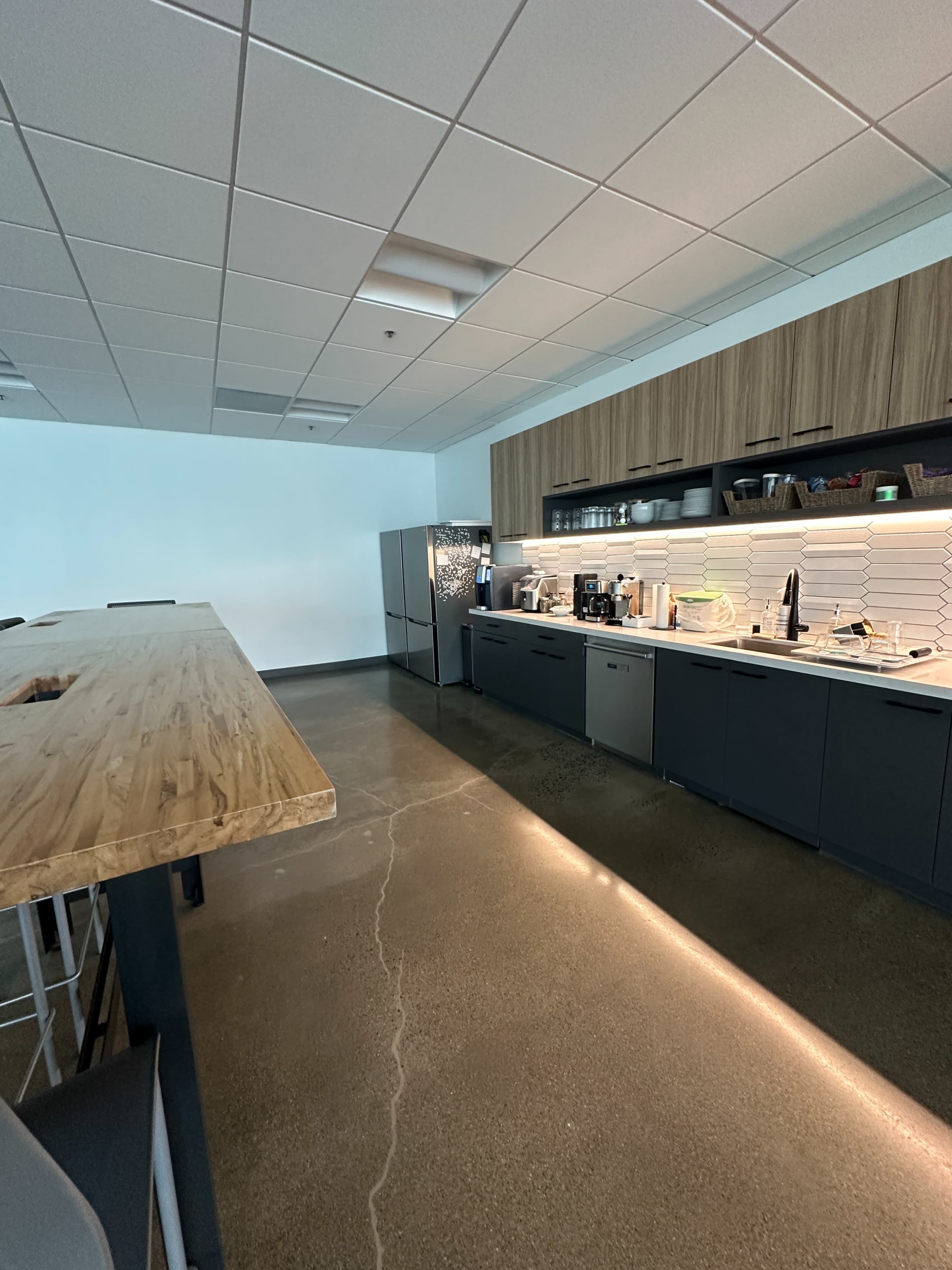 The image shows a modern kitchen area with wood and gray cabinetry, a stainless steel refrigerator, and a long wooden table with barstool seating.