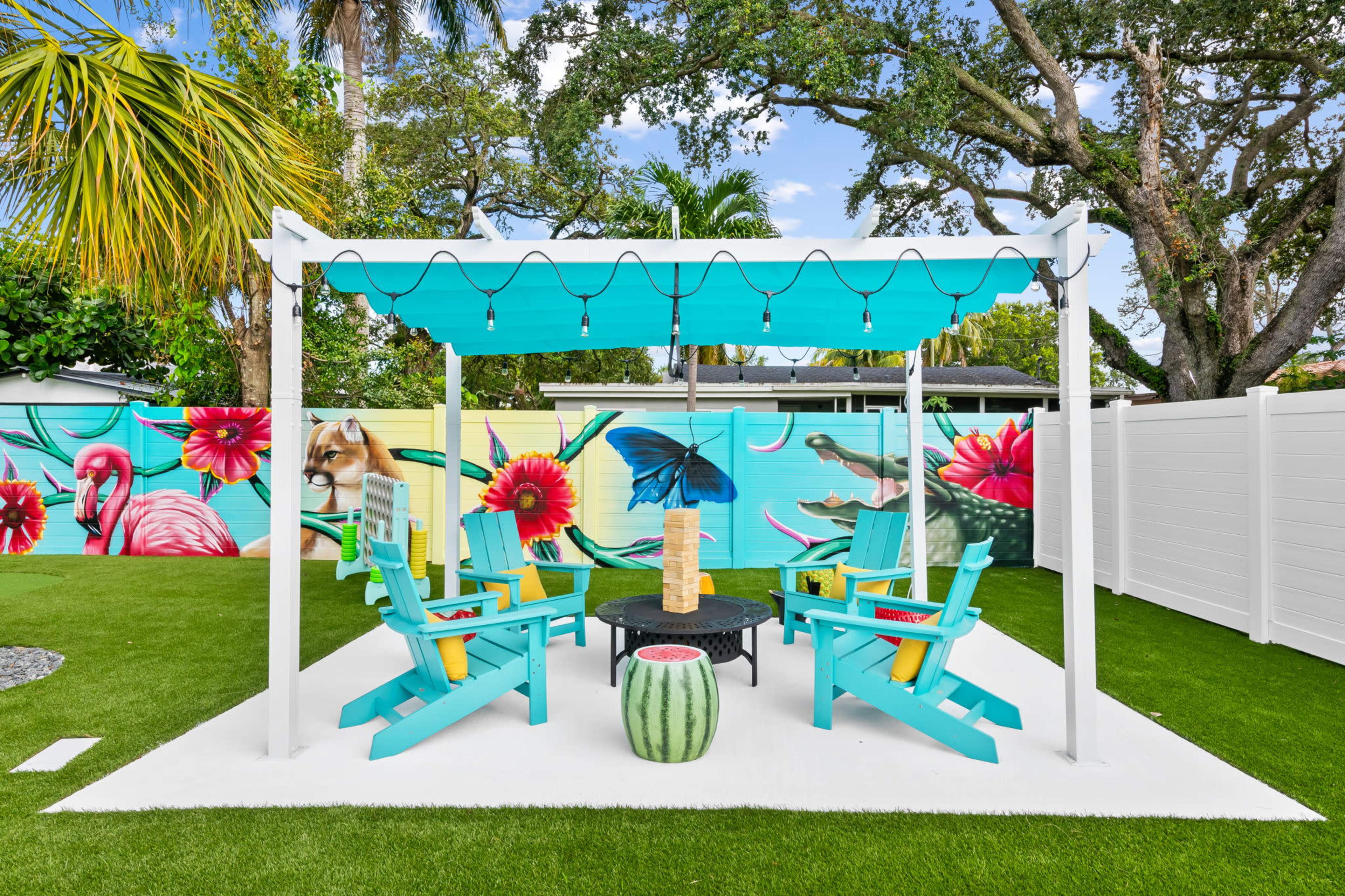 The image features a vibrant outdoor seating area with colorful chairs arranged around a circular table, surrounded by lush greenery and a mural depicting tropical elements.
