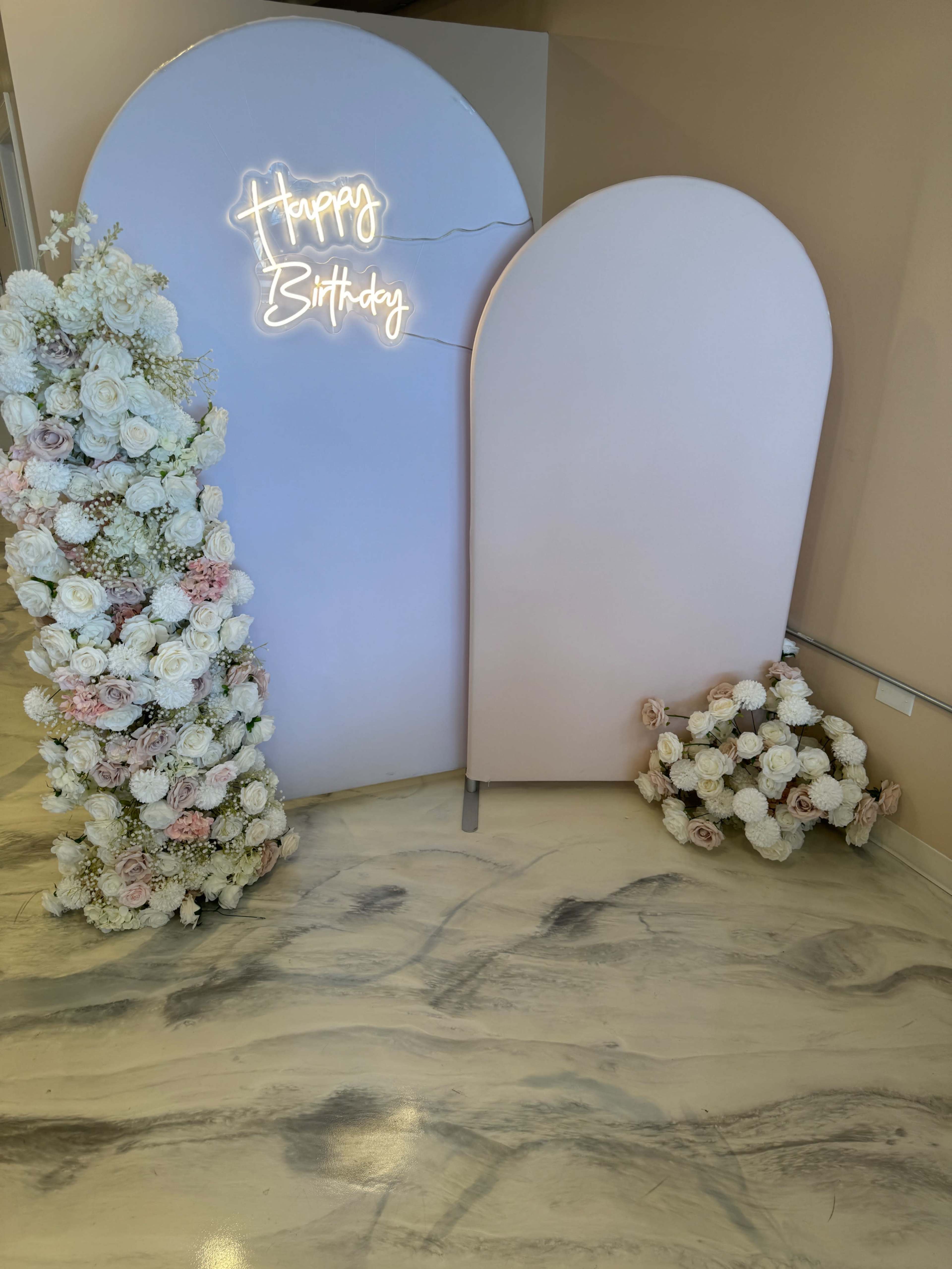 A backdrop with a "Happy Birthday" neon sign, flanked by floral arrangements on one side and a smaller floral decoration on the floor.