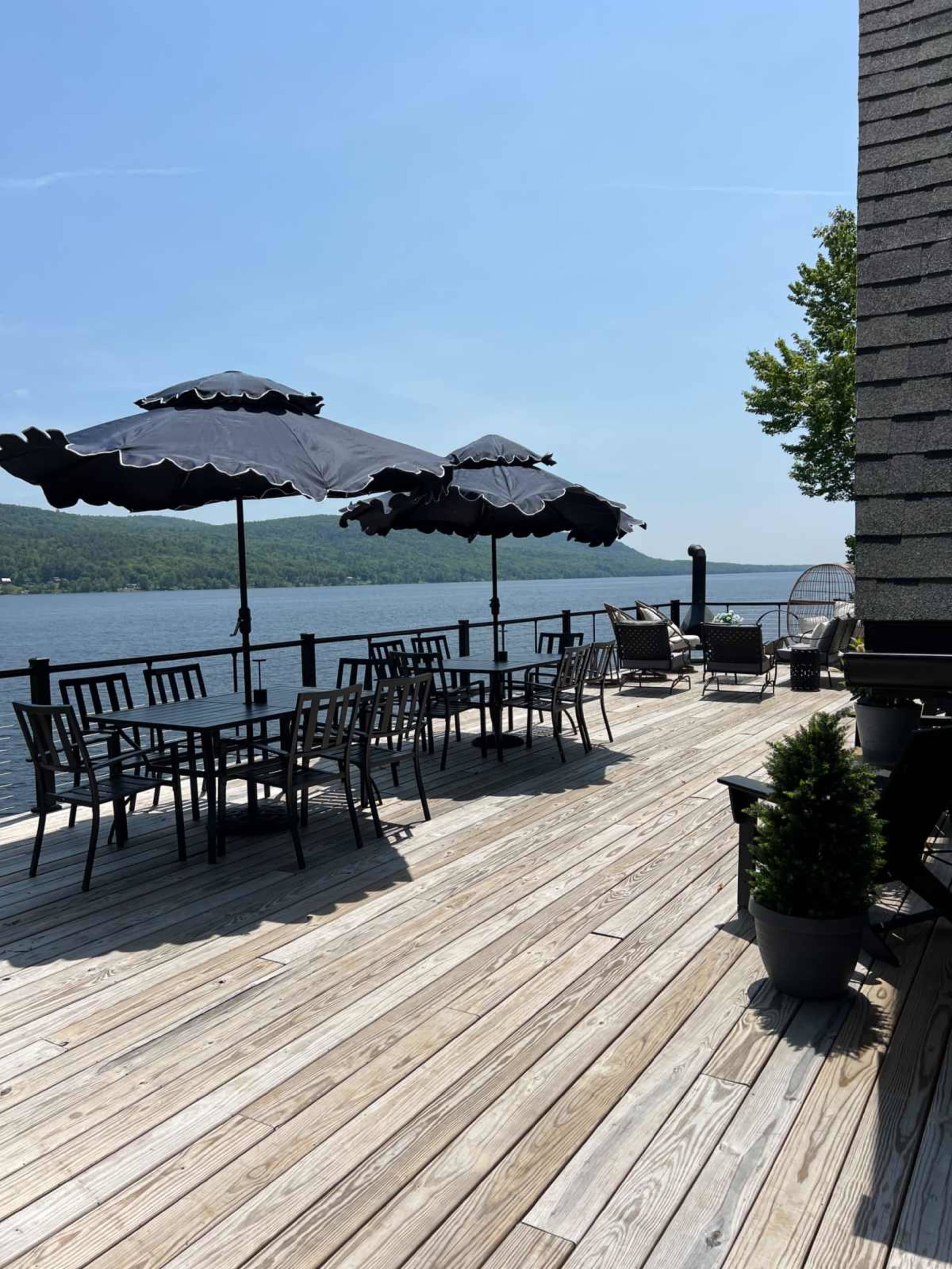 A wooden deck overlooks a calm body of water, featuring several tables with black umbrellas and seating arrangements.