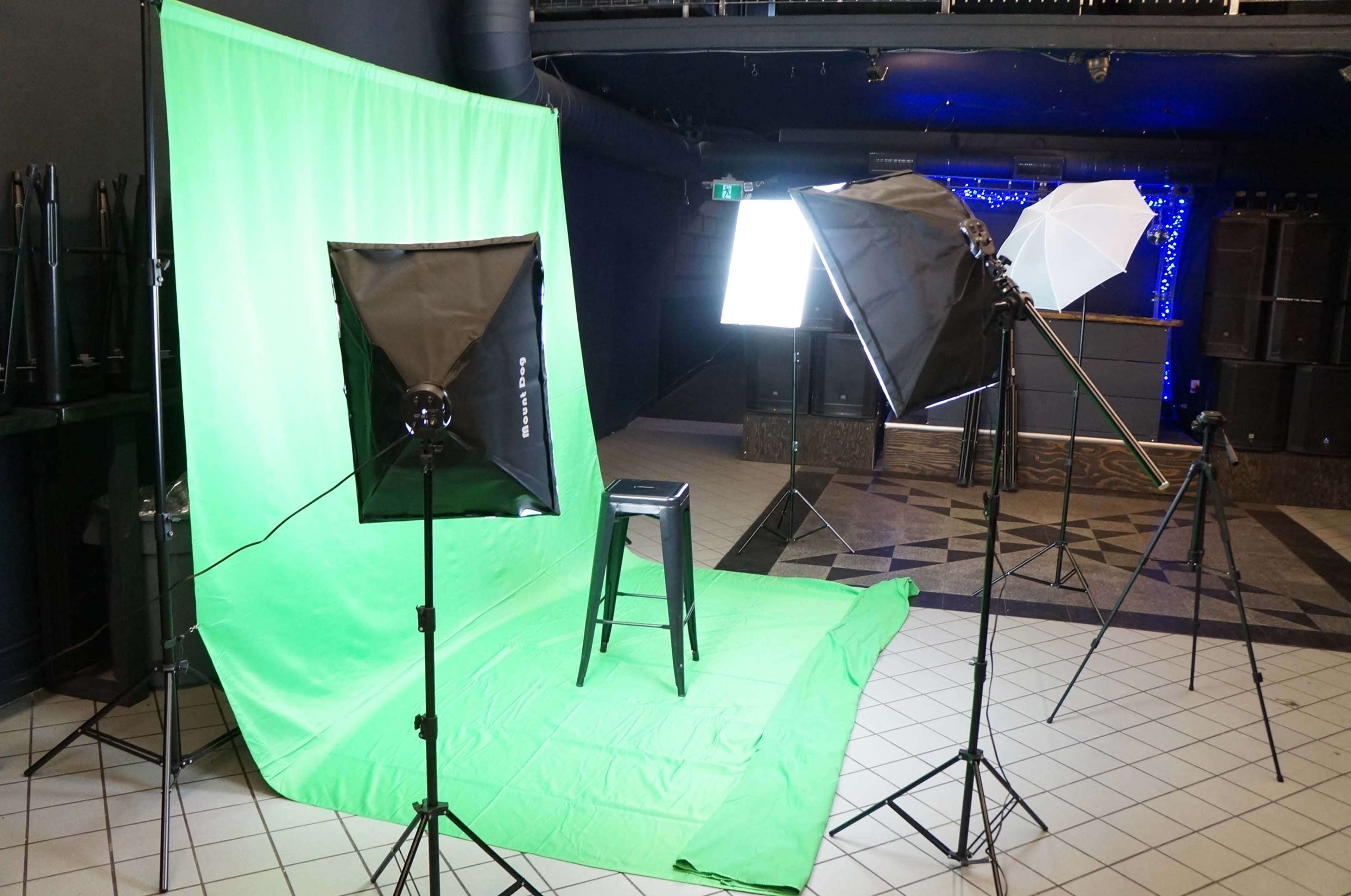A photography setup featuring a green backdrop, a black stool, and several studio lights arranged on tripods in a spacious room with a patterned floor.