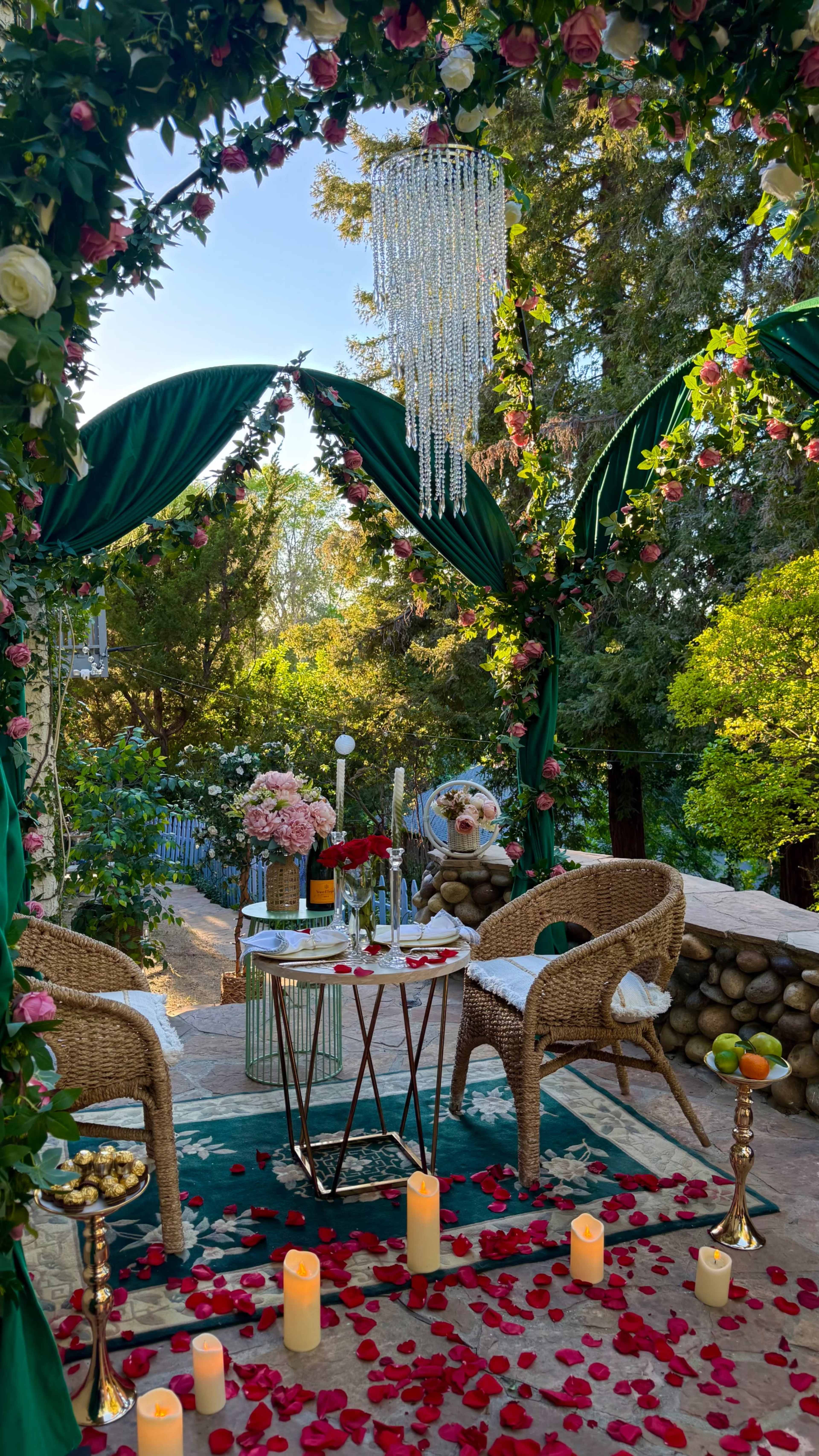 A cozy outdoor seating area is adorned with greenery and flowers, featuring two wicker chairs, a small table set with refreshments, and a crystal chandelier hanging above.