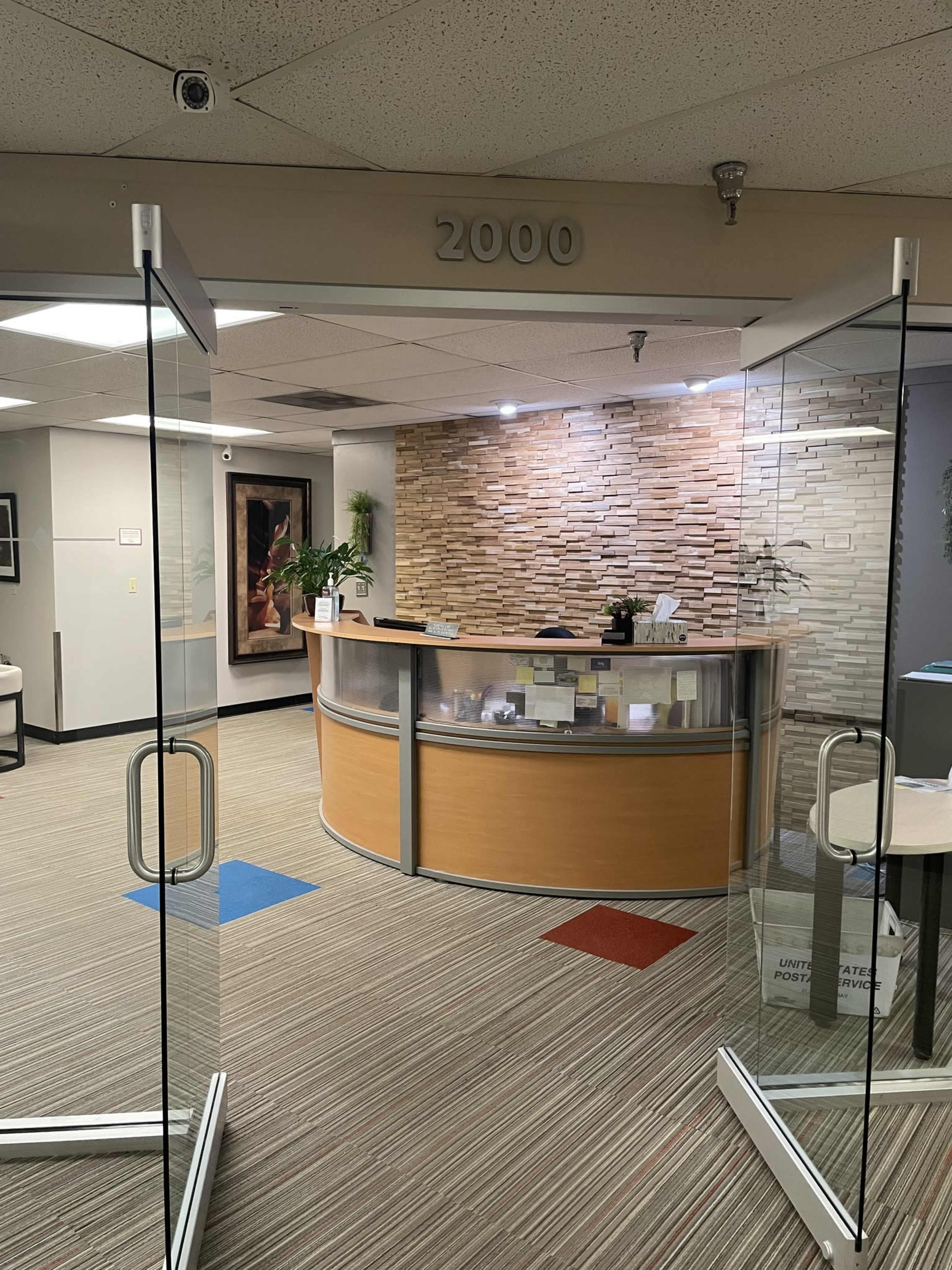 The image shows a reception area with a curved front desk, a stone wall backdrop, and a clear glass entrance.