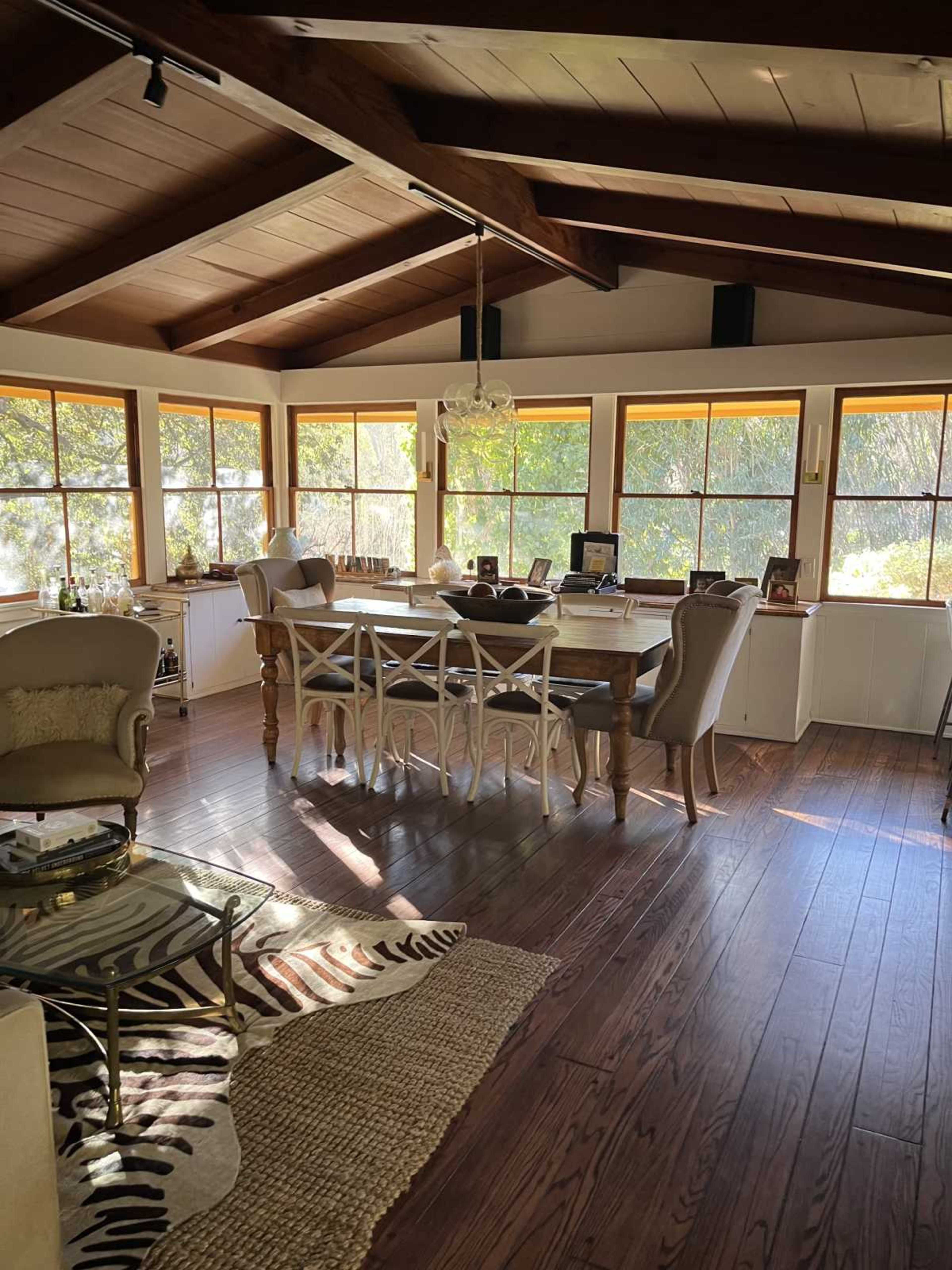 The image shows a spacious, well-lit interior with a wooden ceiling, a dining table surrounded by chairs, and large windows overlooking a garden.