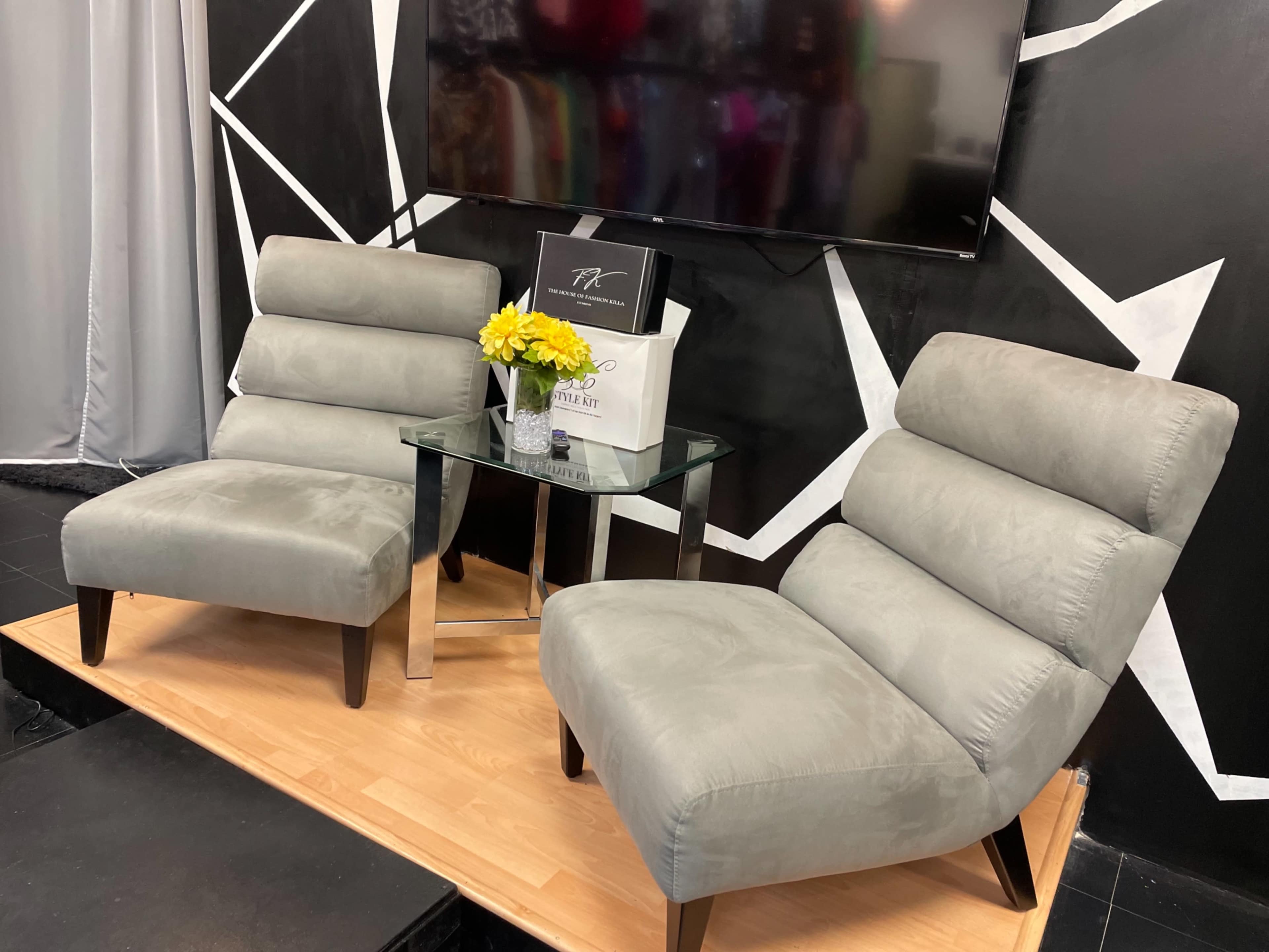 The image shows two gray upholstered chairs placed beside a glass table with a small bouquet of yellow flowers on it, set against a black and white geometric backdrop.