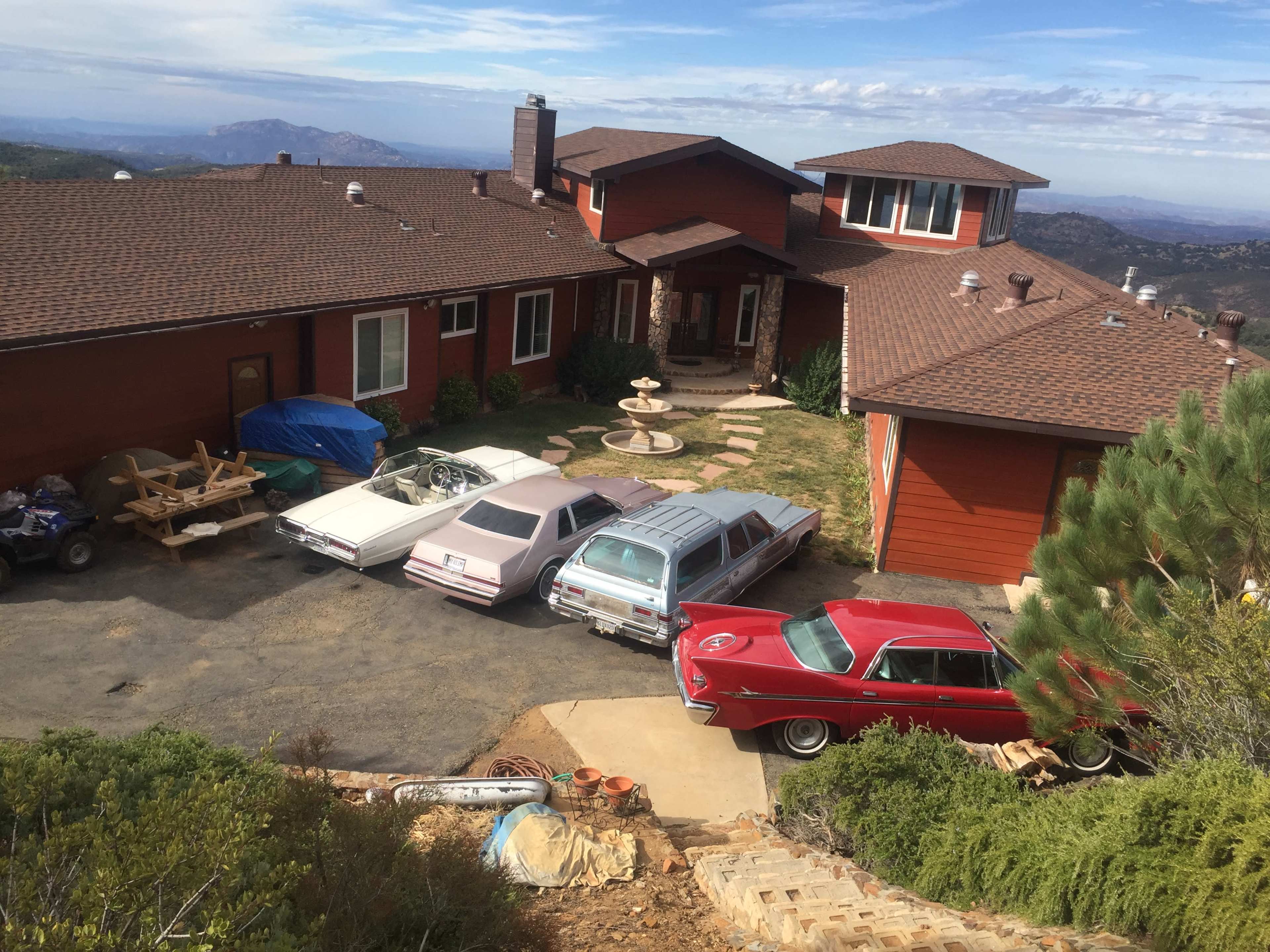A red car and two vintage cars are parked in front of a large brown house set on a hillside, with mountains visible in the background.