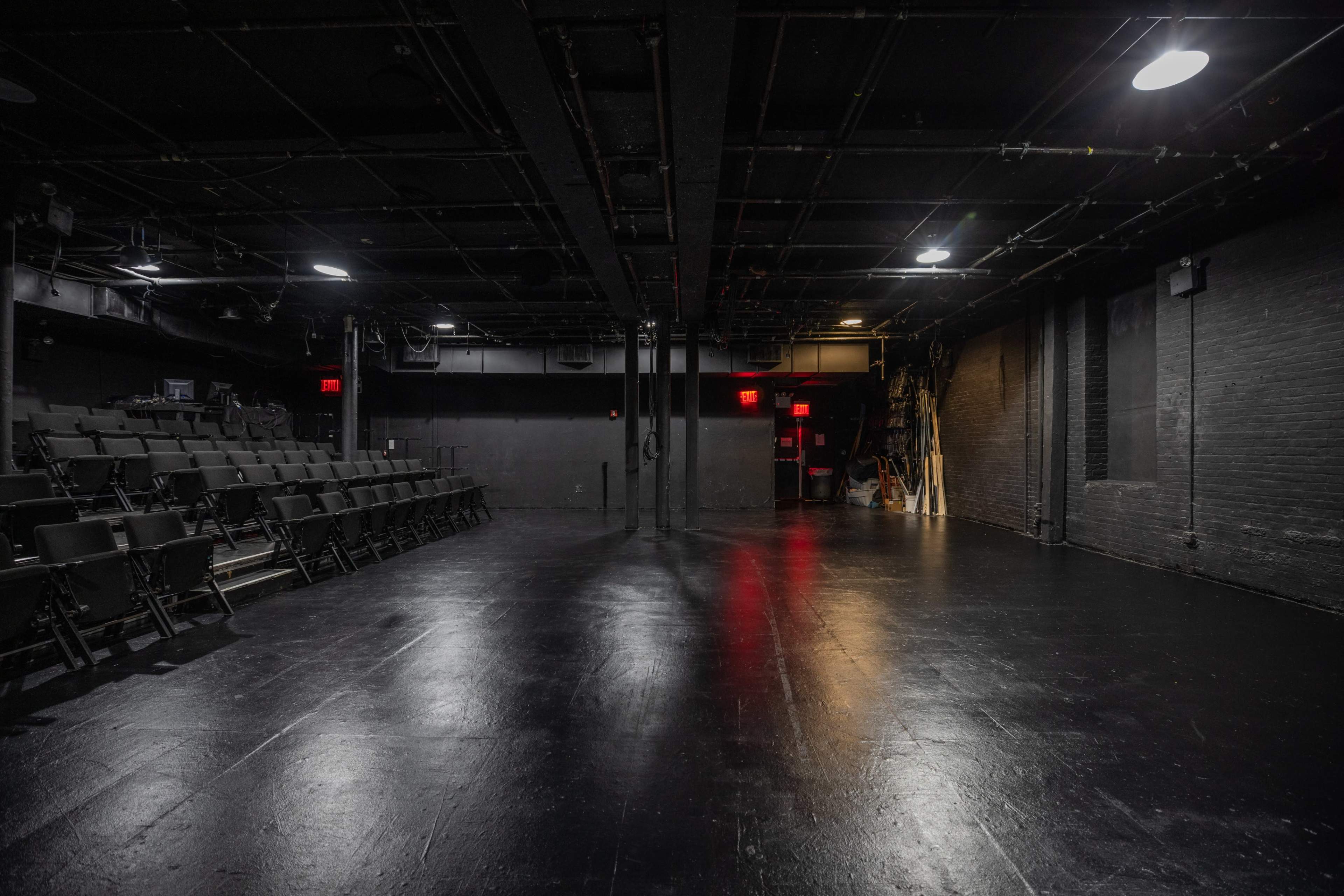 The image shows a black-box theater with empty rows of seats on one side and a dark stage area lit by overhead lights.