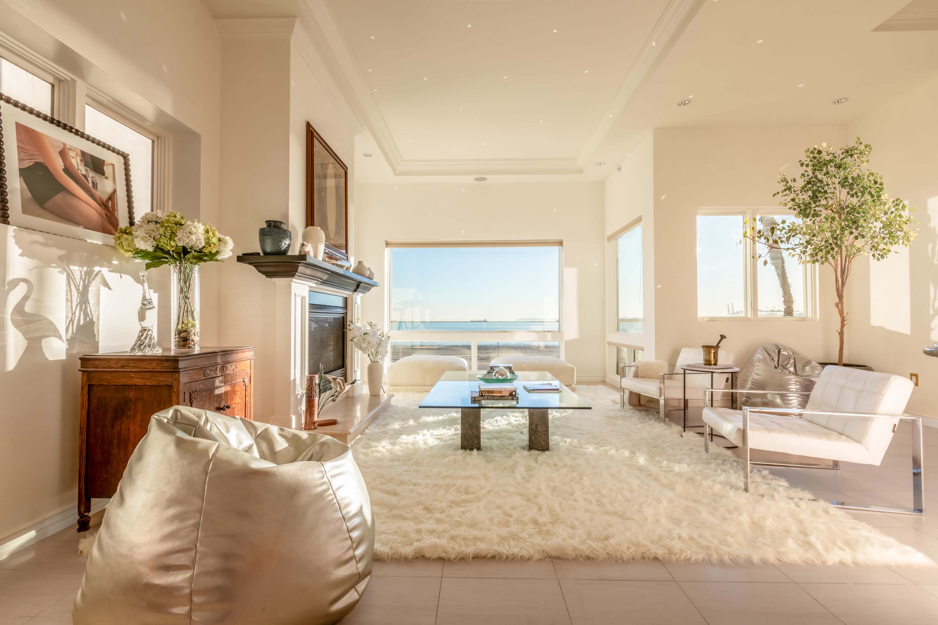 The living room features a large window with a view of the water, a fireplace, and modern furniture arranged on a fluffy white rug.