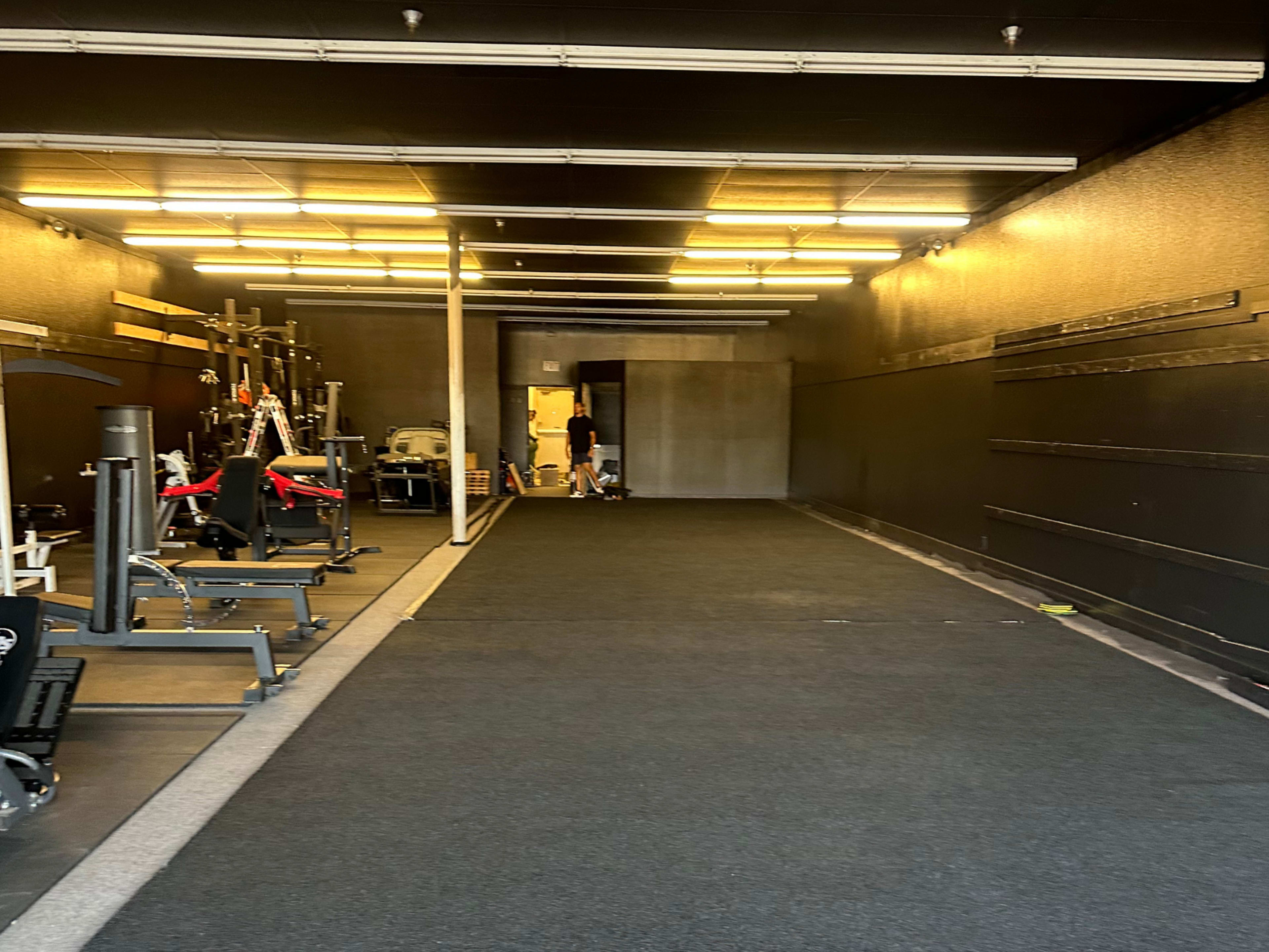 The image shows a dimly lit gym interior with workout equipment on one side and an open hallway leading to another area.